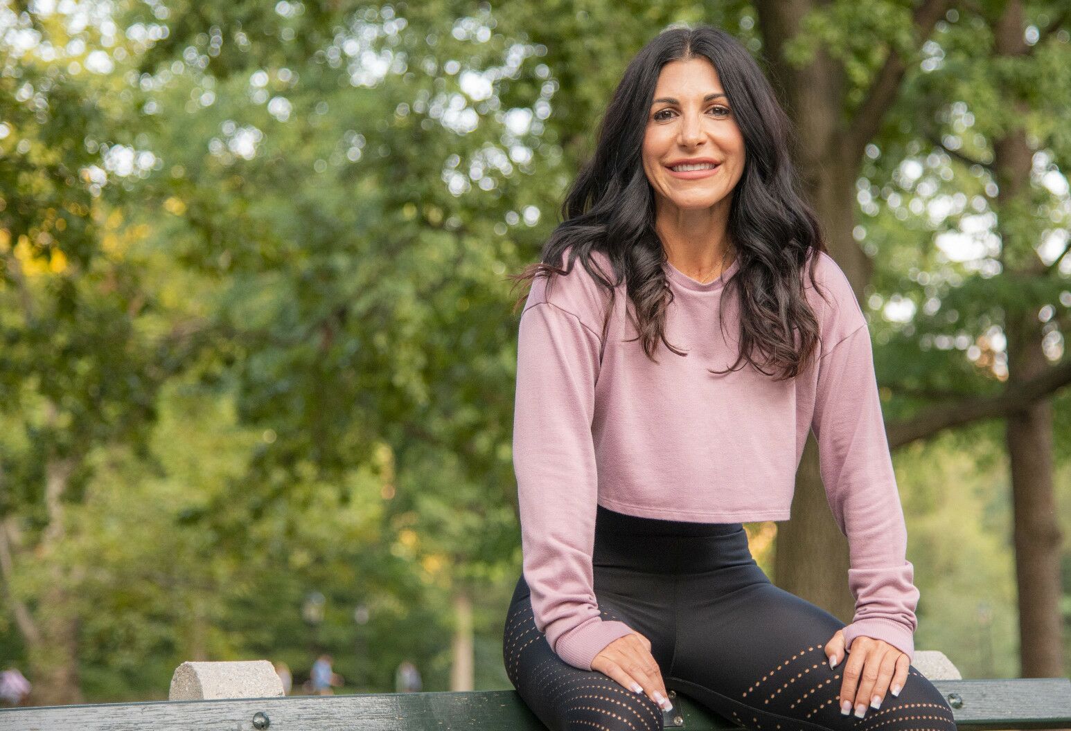 Woman with long, brown hair and pink long sleeve shirt sits on the back of a park bench.