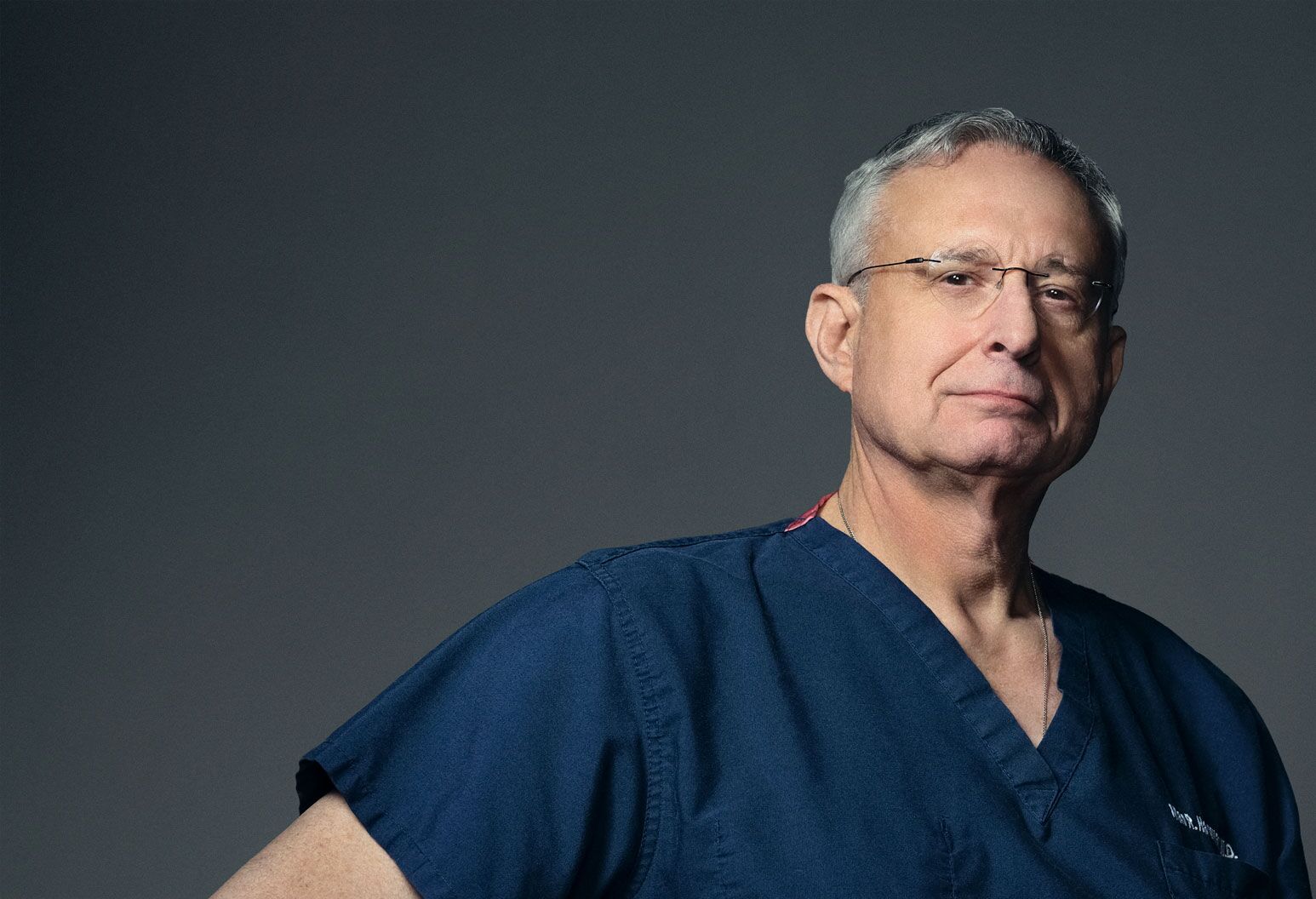 Physician in blue scrubs looking straight ahead against a dark background.