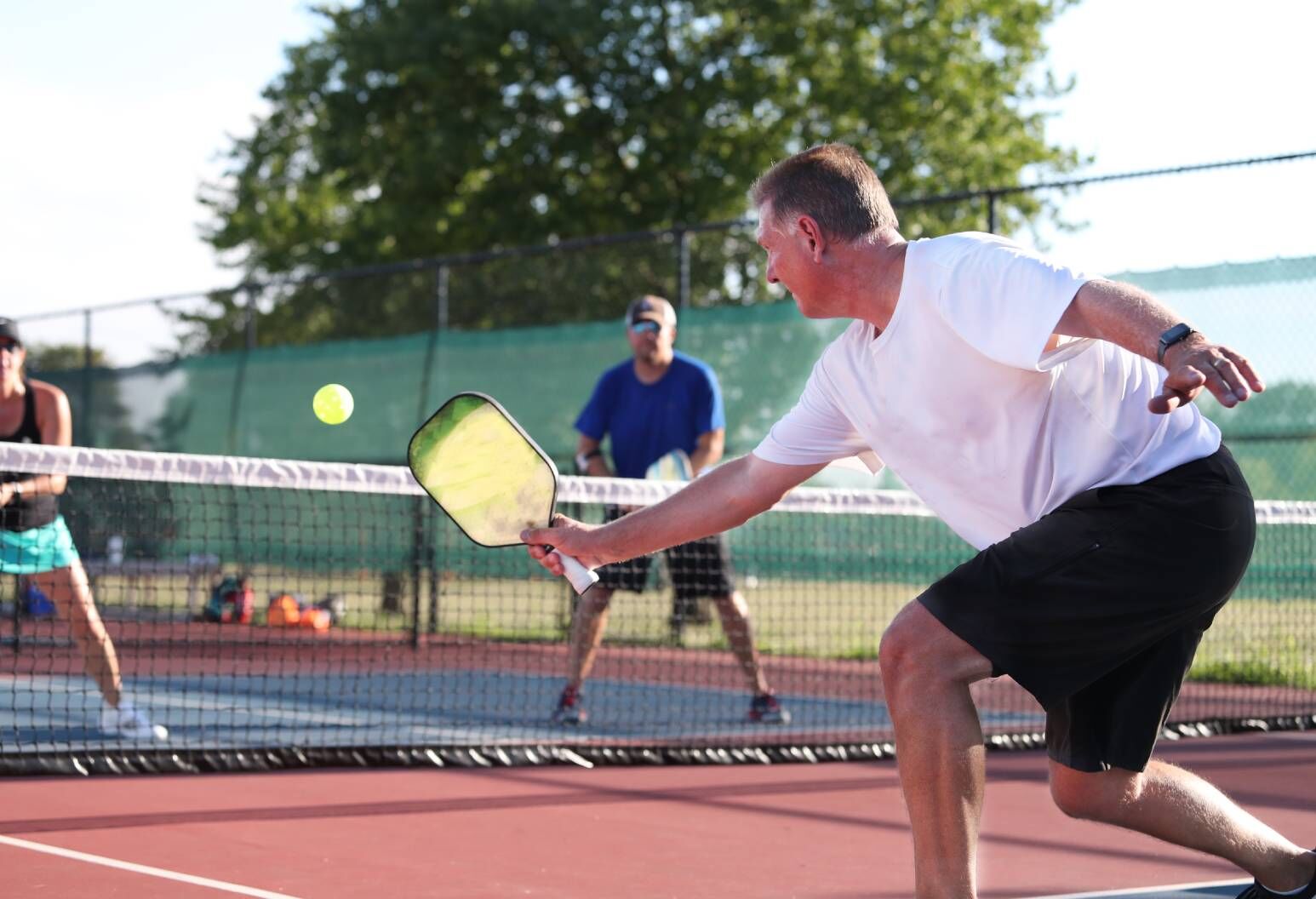 A middle-aged man in activewear hits a pickleball toward the net of a tennis court, with two opponents standing in the background, on the other side of the net.
