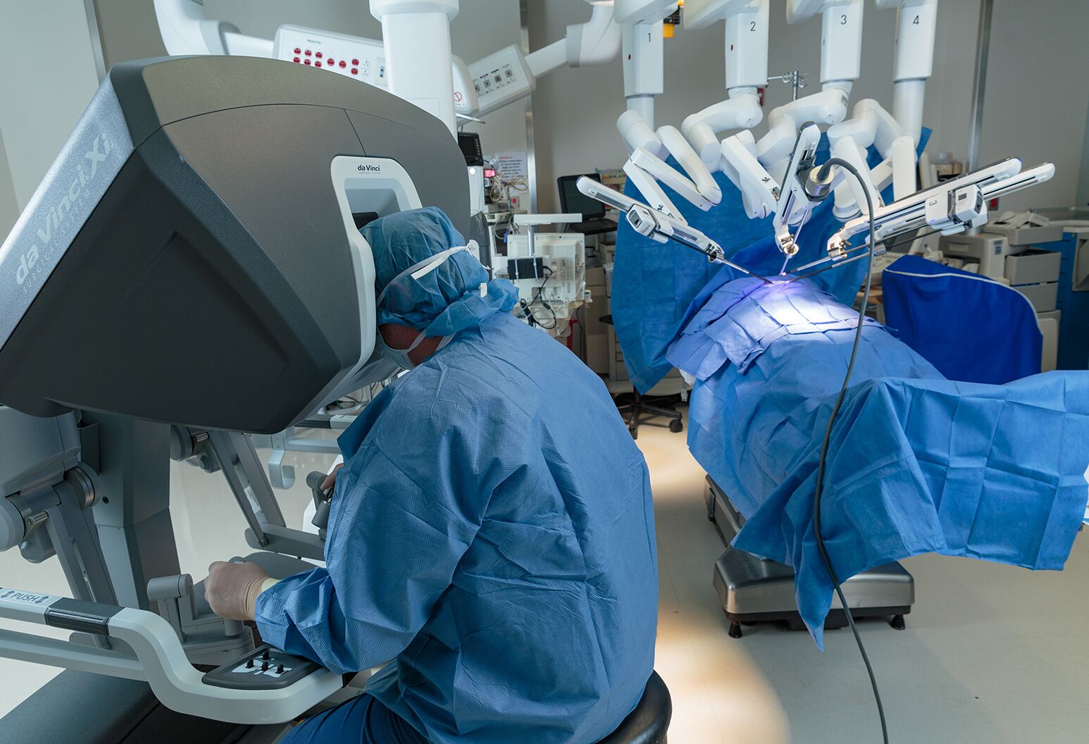 A surgeon in foreground performs robotic surgery on a patient under a sheet in background.