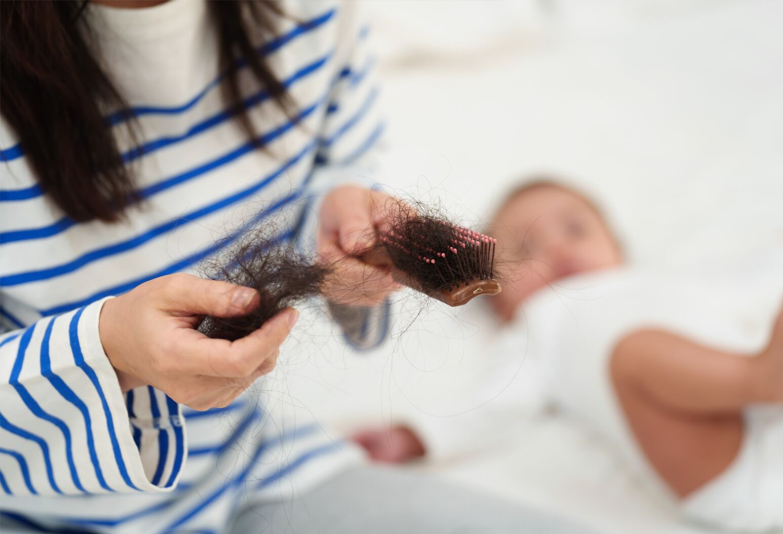 A woman holding a hair brush filled with hair, with a baby lying next to her.