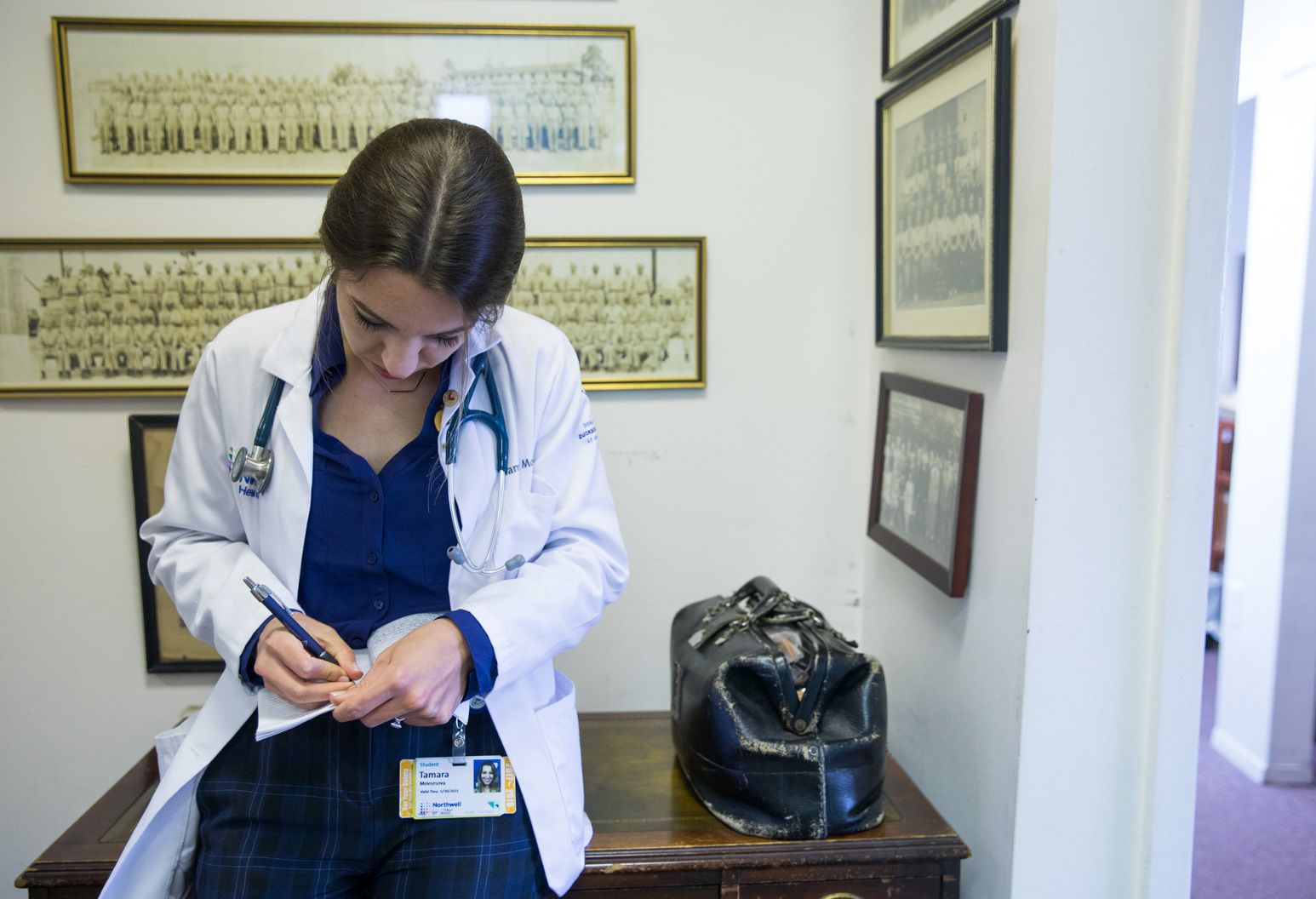 A woman in a blue blouse wearing a white doctor's coat and a stethoscope hangs around her neck as she leans against a table writing in her notebook. Next to her on the table is an aged black leather doctor's supply bag. On the walls are portraits of old black and white photos filled with group photos of people in uniform.