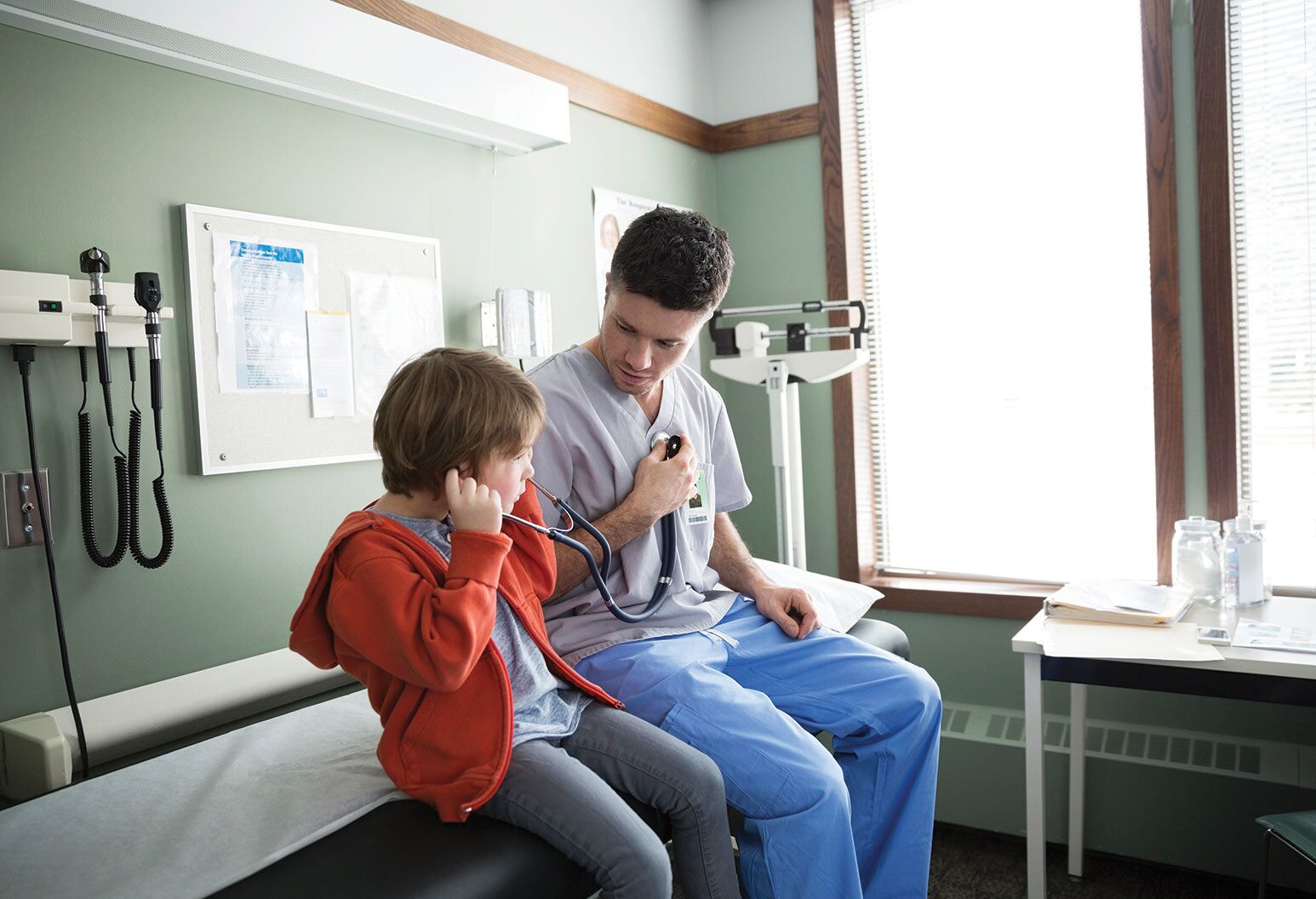 A provider sits next a young boy on the hospital bed, letting him use his stethoscope.