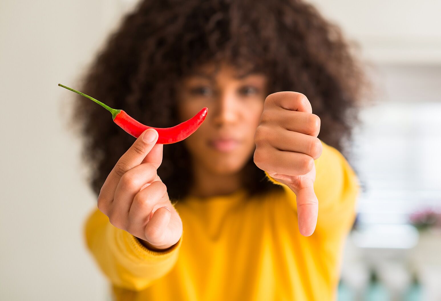 A woman in an orange sweater holding a red pepper with one hand and giving a thumbs down with the other.