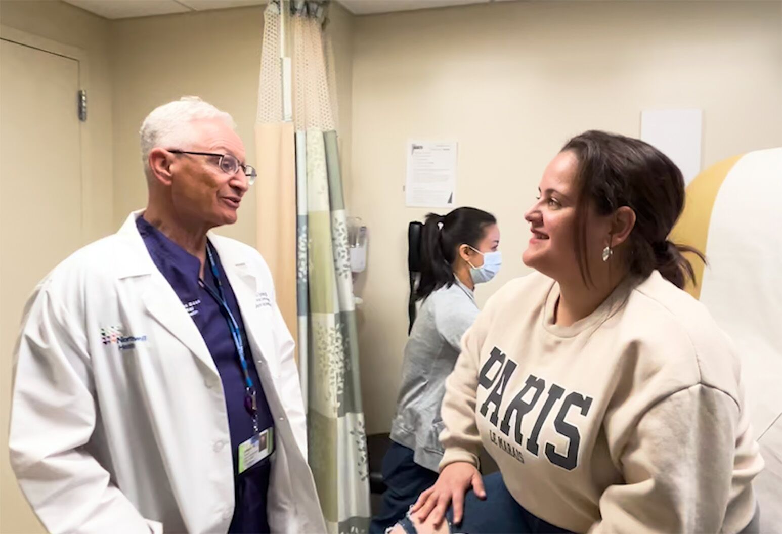 A male doctor wearing a white lab coat speaks with a young female patient sitting on a doctor's office chair. A female health care worker works on a computer in the background. 
