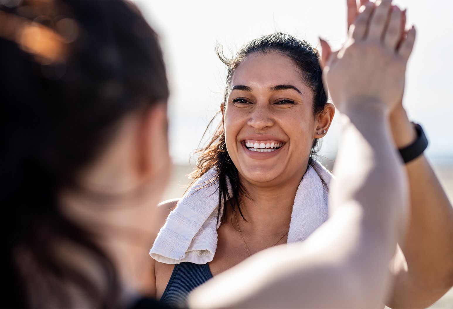 Smiling woman with dark brown hair and white towel around her neck gives her friend a high five after exercising.