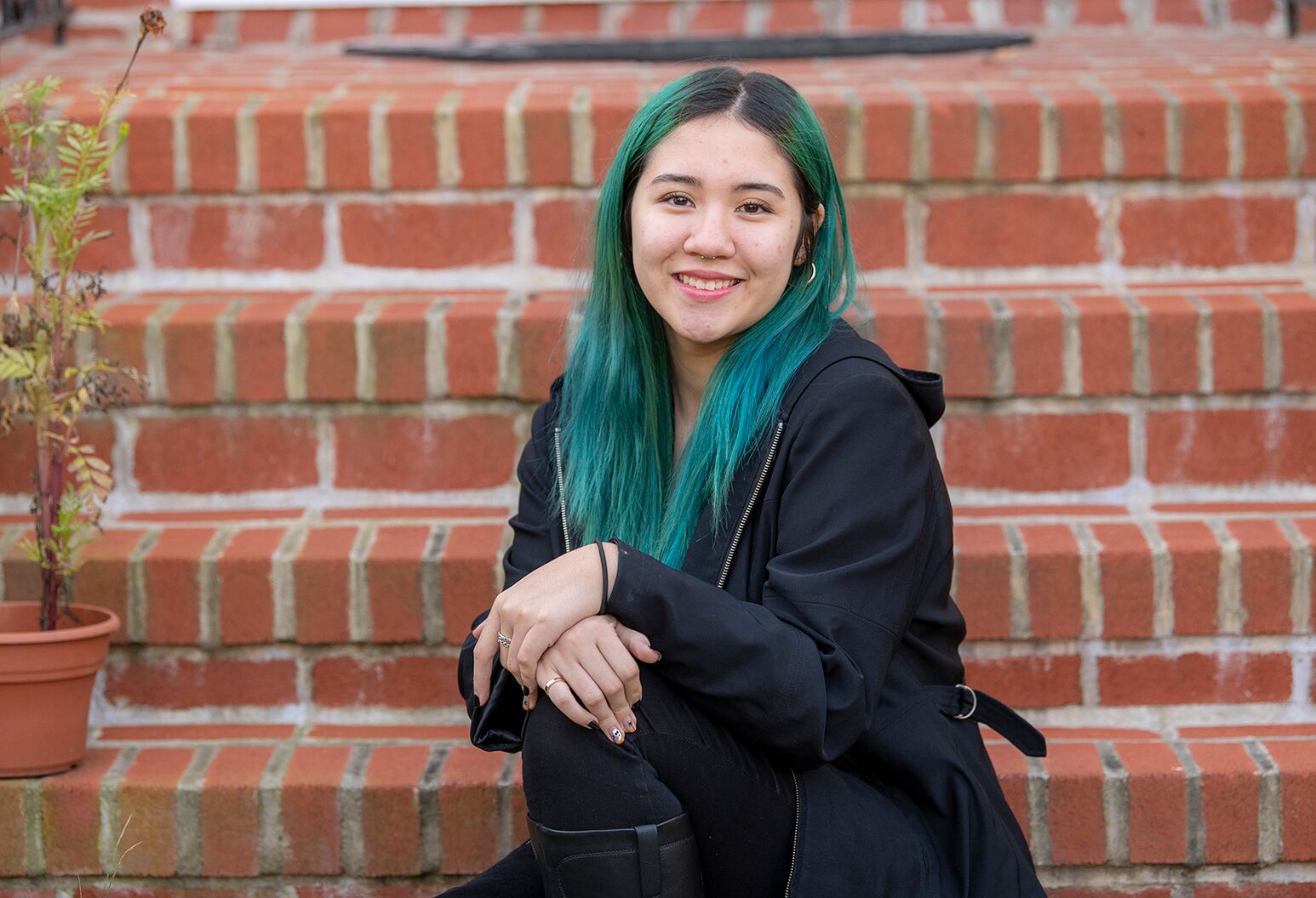 Young smiling woman with dyed green hair and black jacket on a brick porch.