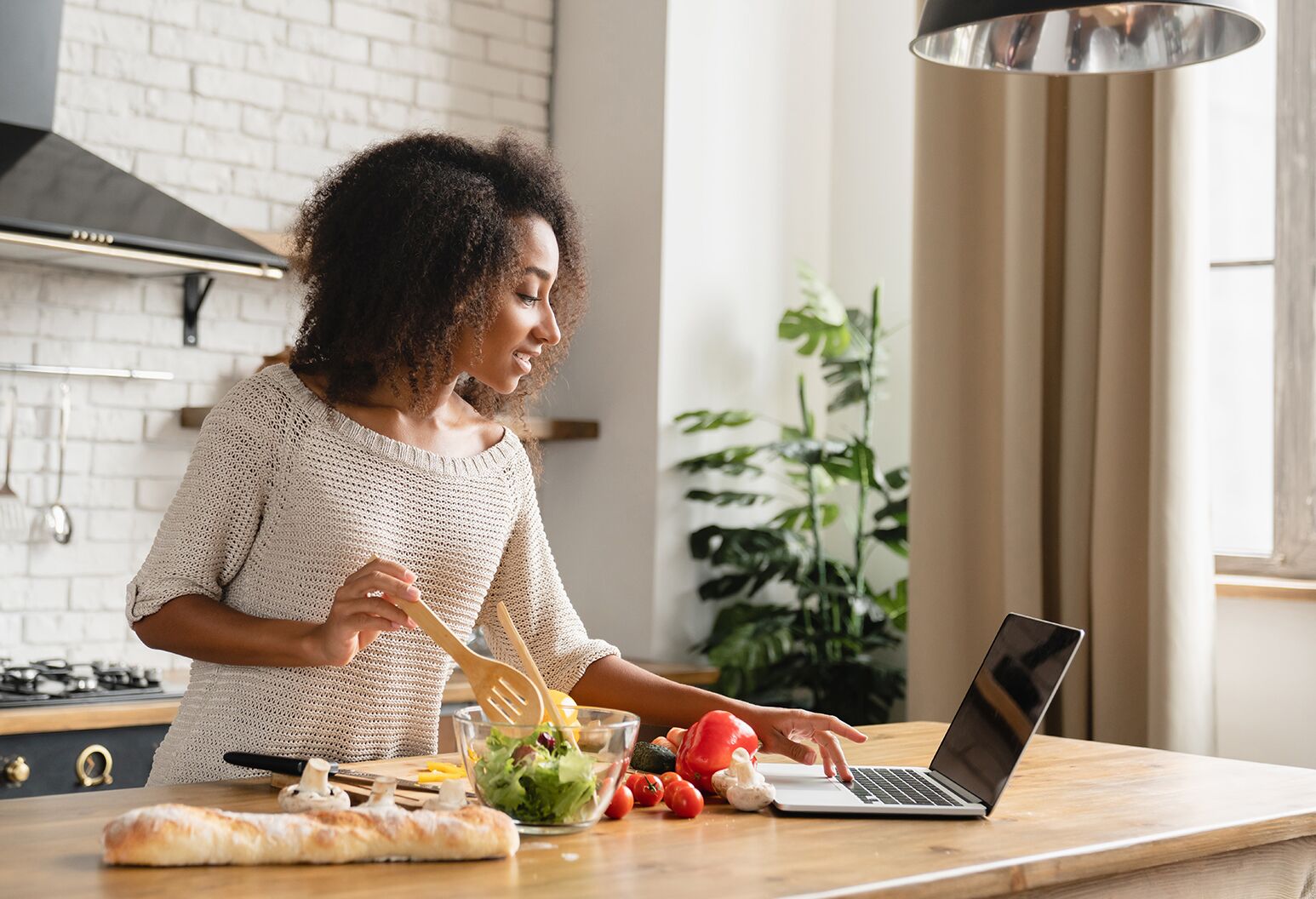 A woman in a kitchen overlooks several vegetables on a cutting board and looks at her laptop.