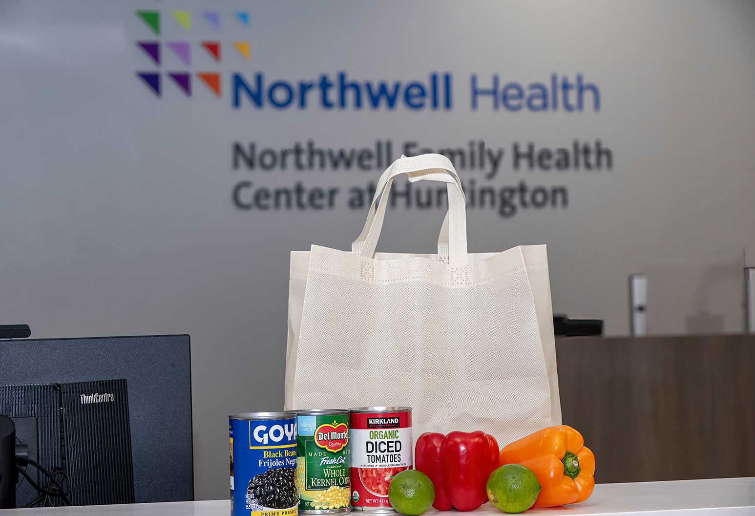 Cans of food and loos vegetables surround a white tote bag on a table, with signage on the wall behind it that reads "Northwell Health: Northwell Family Health Center at Huntington."