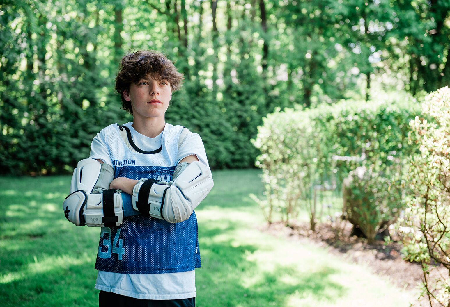 A teenage boy in lacrosse gear crosses his arms and looks to his left in a wooded scene.