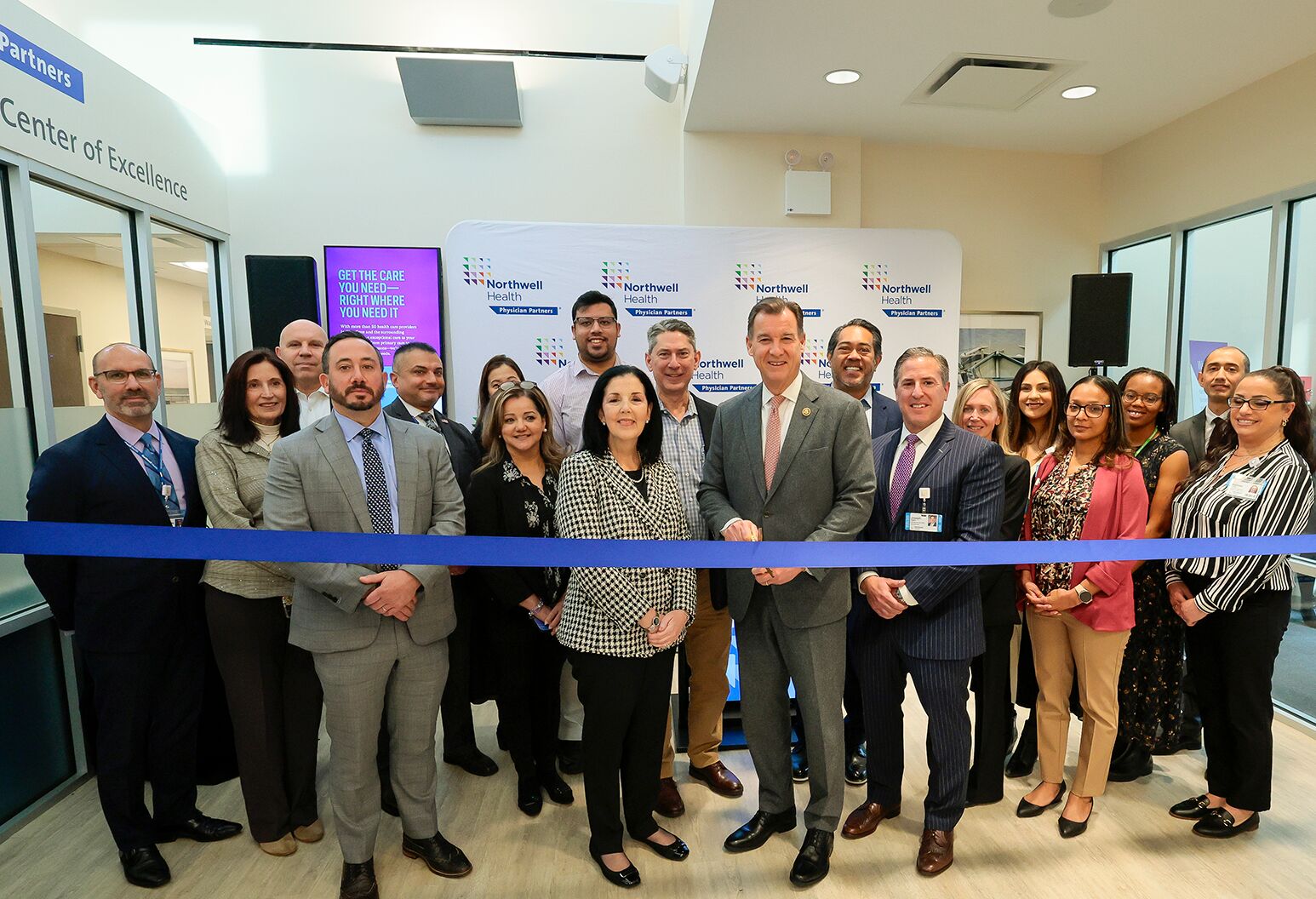 A group of professionally-dressed men and women stand behind a blue ribbon in a medical building. 