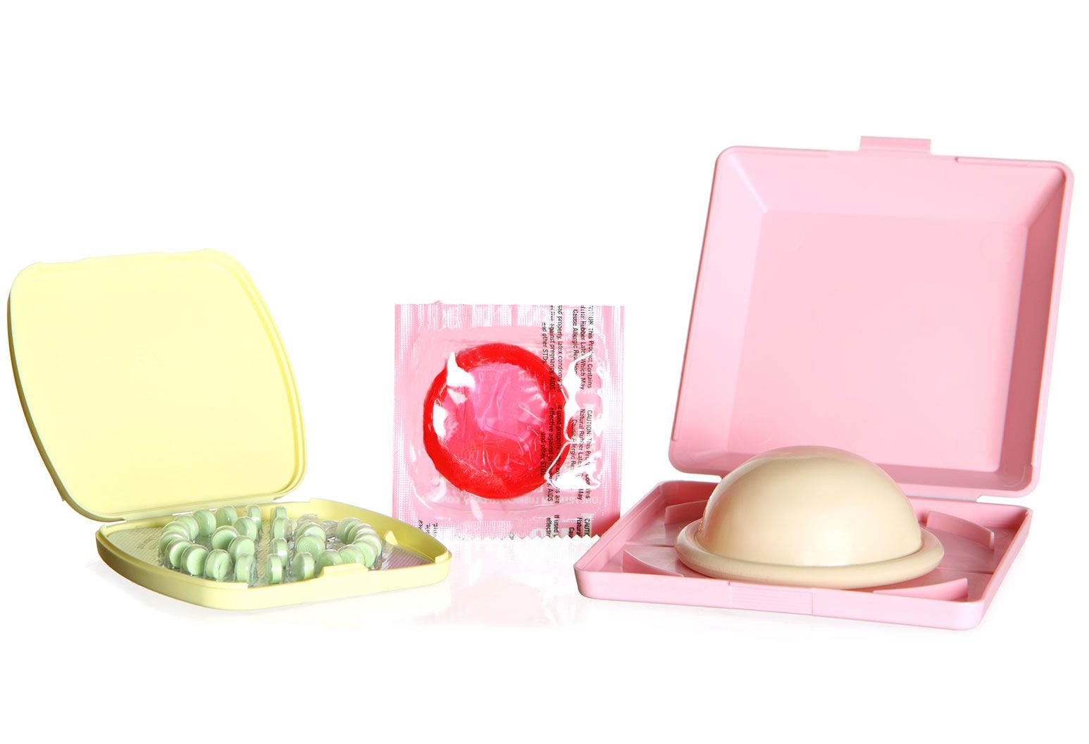 A yellow pill box containing green pills, an upright red latex condom in packaging, and a pink box holding a diaphragm are positioned next to each other.