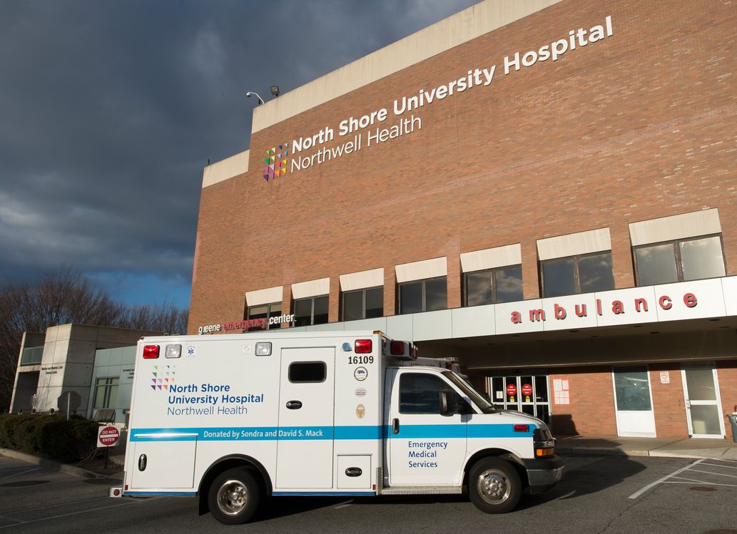 White ambulance with blue "North Shore University Hospital" writing is parked outside a brown, brick building with the words "North Shore University Hospital"