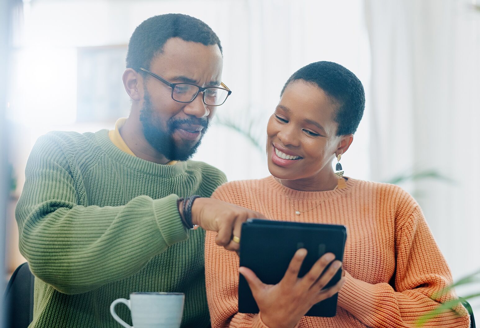A man wearing a green sweater sits next to a woman in an orange sweater and points over her shoulder at a tablet she holds.