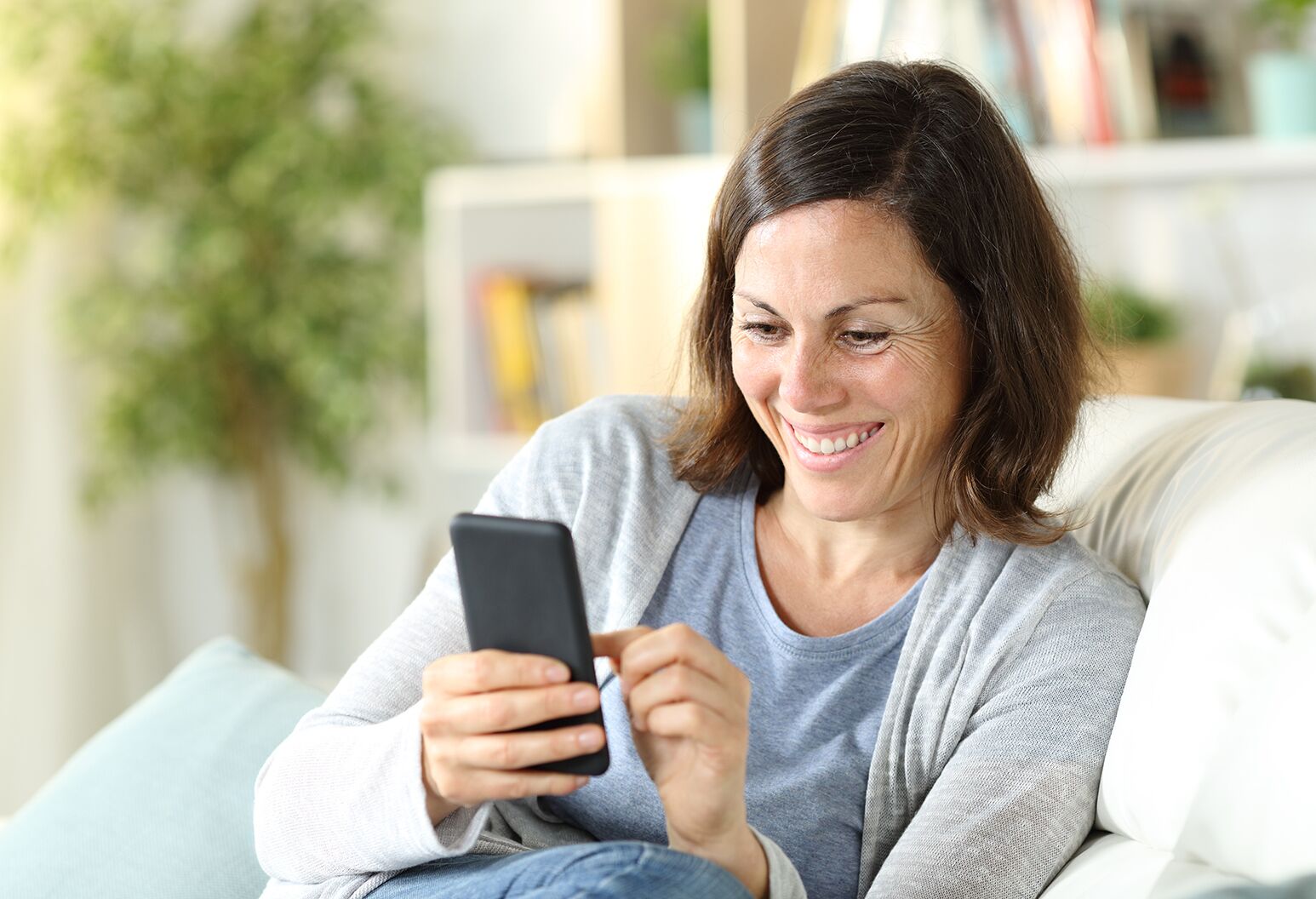 A young woman with brown straight hair is using mobile phone. Female is smiling while holding smart phone. She is sitting on sofa at home.