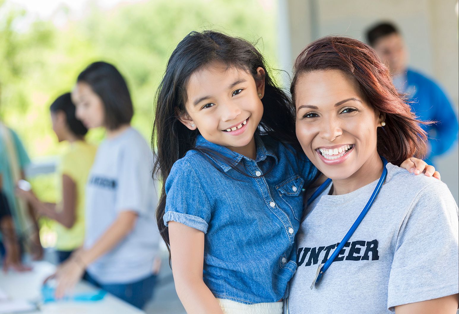 Cheerful volunteer wearing a grey t-shirt that says "volunteer". She is holding on to the little girl. They are blissfully smiling at the camera. There are others working blurry behind them. 