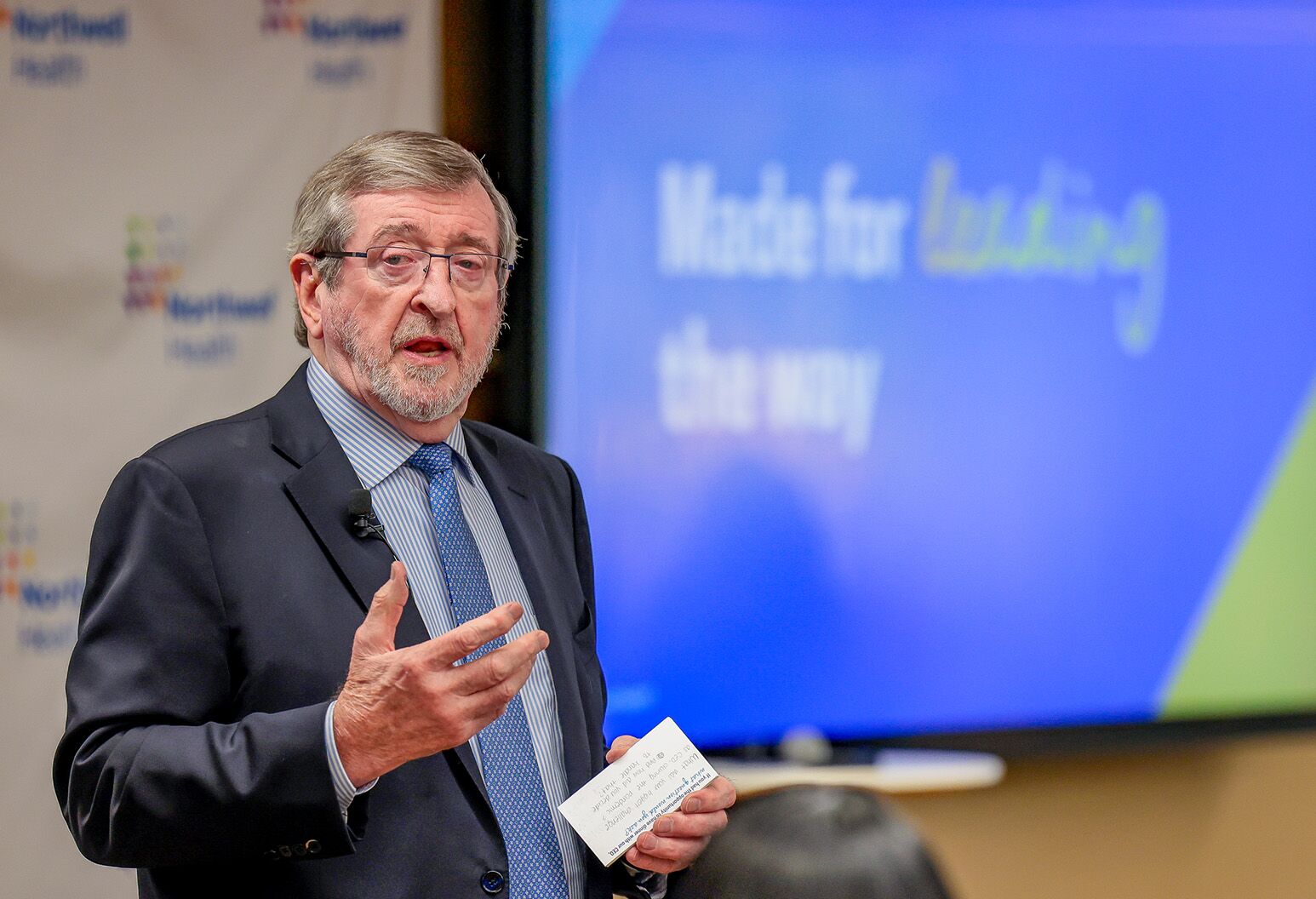 A man in a suit stands speaking to a crowd (not shown) with a video board behind him showing a blue and green slide with white letters that say "Made for leading the way."