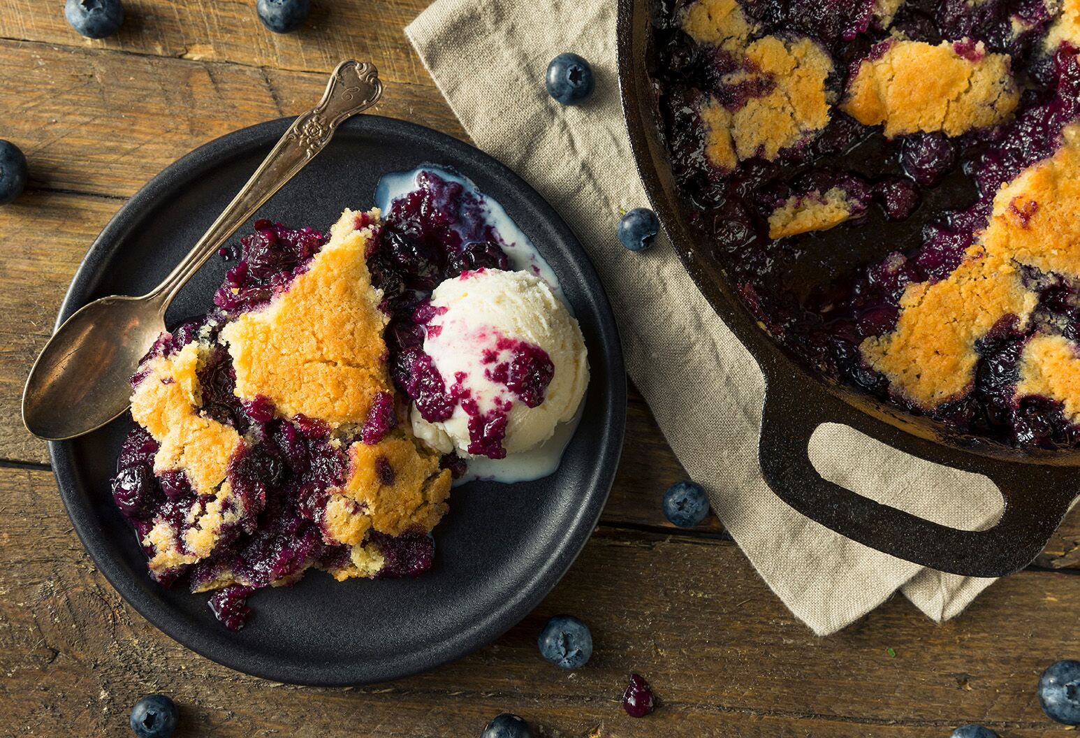 Two white ceramic bowls of blackberry cobbler are pictured sitting atop a black metal cooling rack surrounded by two spoons and cuttings of blackberry vines.