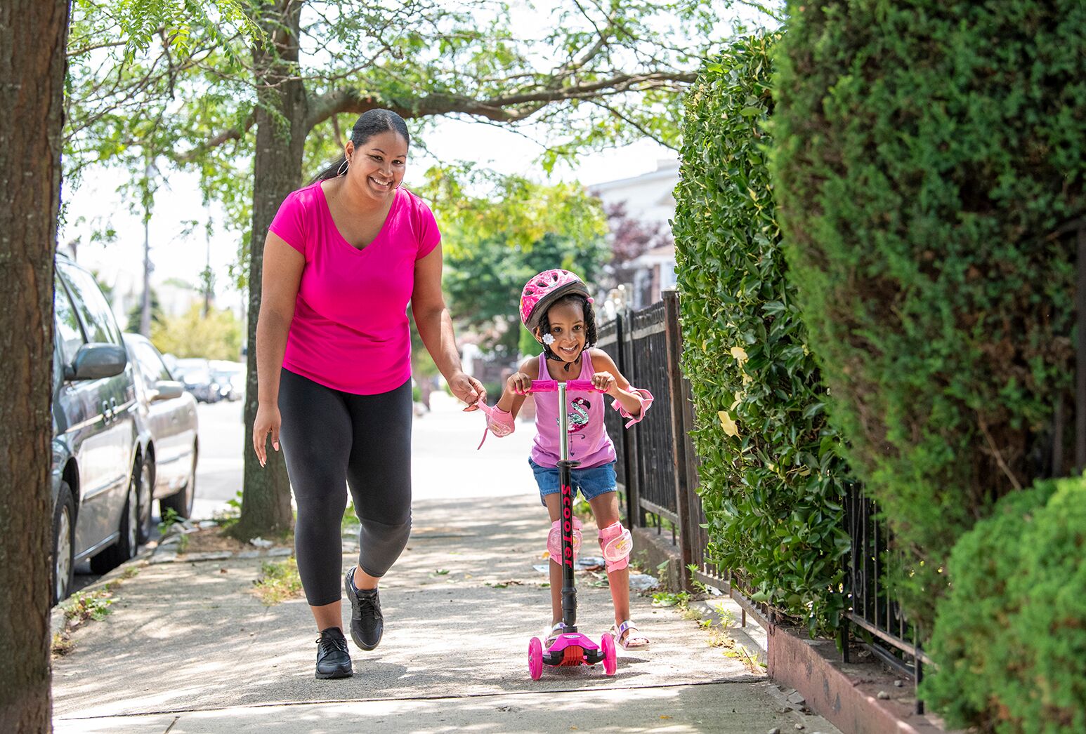 African American woman in fuchsia top and little girl on a pink scooter on the sidewalk.