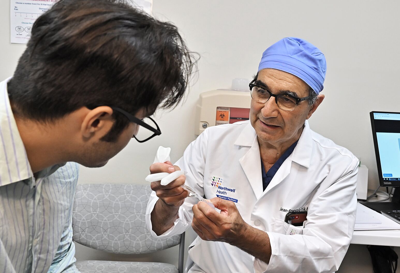 A provider in a white lab coat and surgical scrub cap seated with a patient, talking.