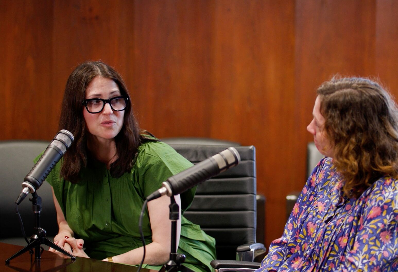 Two women sit at a table and talk into microphones against a wood backdrop.