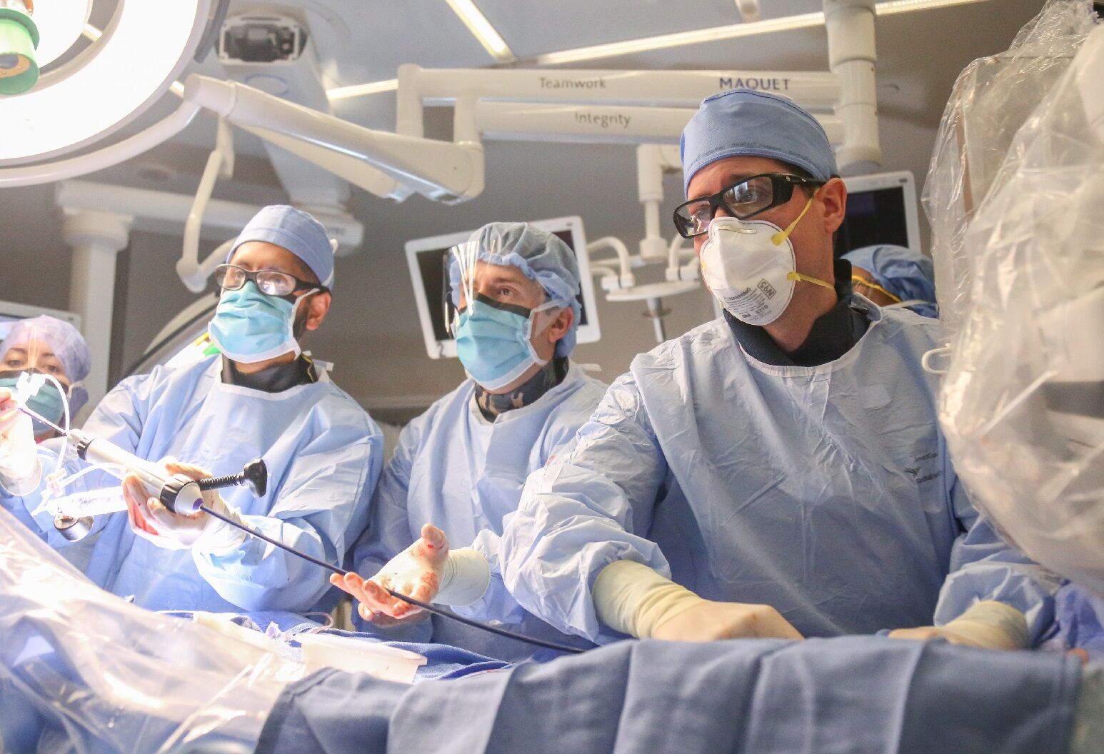 A group of three surgeons in full surgical attire standing over a patient look over at something out of frame.