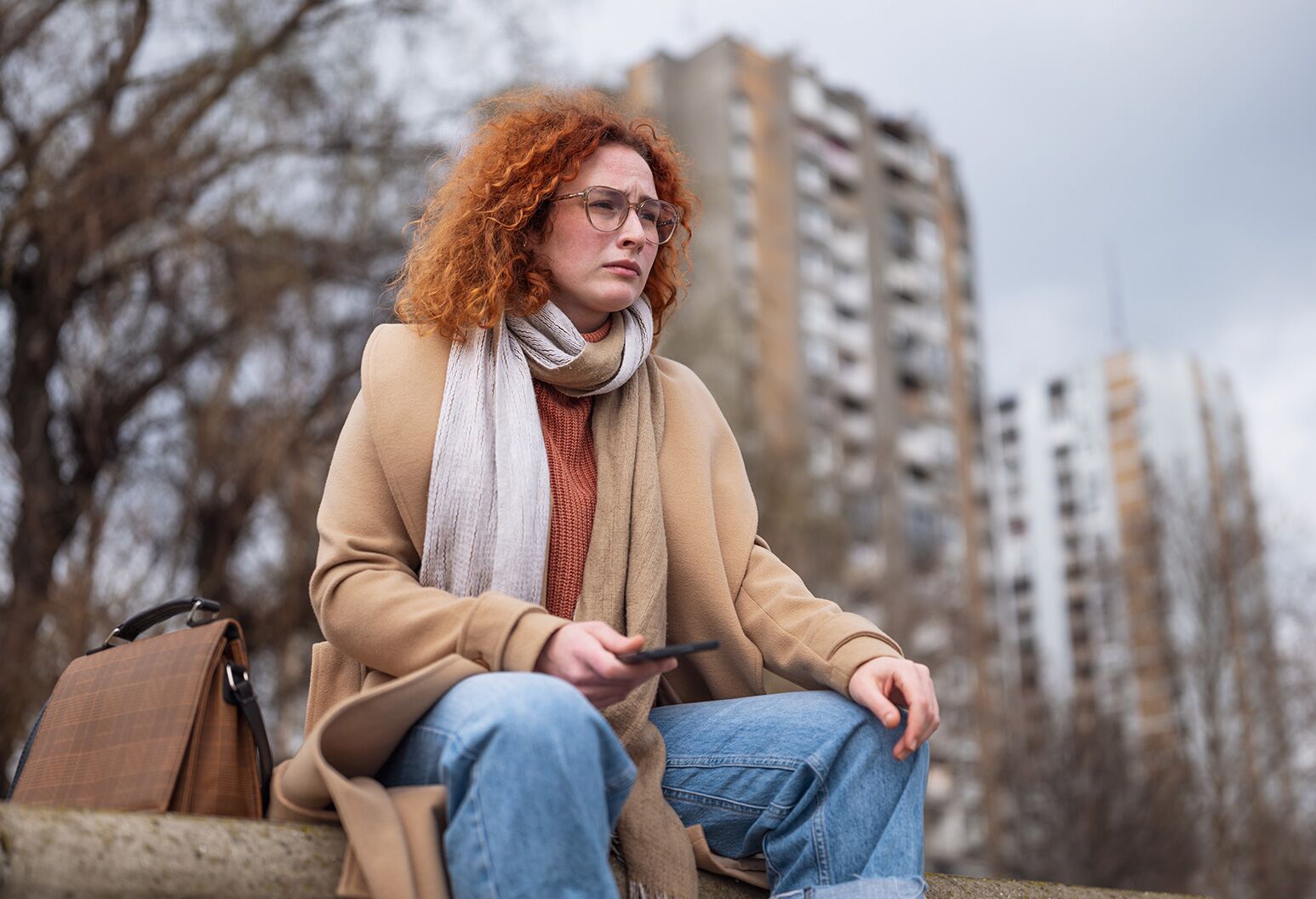 A solemn woman sitting outside on a winter day, holding her phone and looking off in the distance.