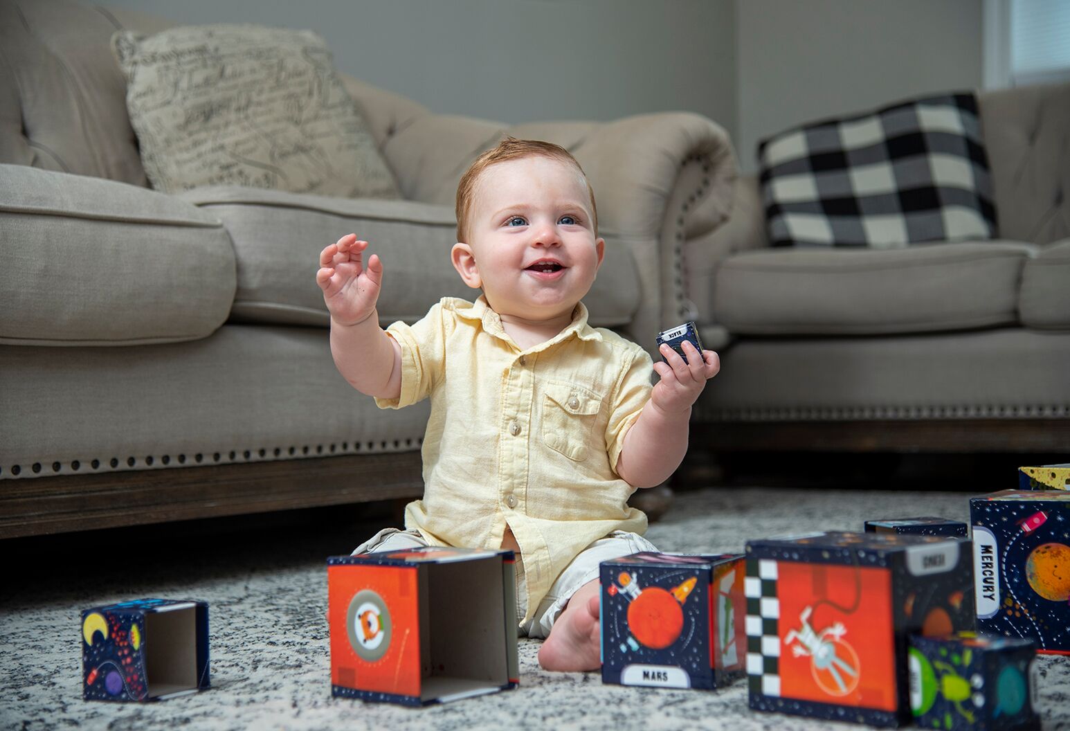 Baby boy with red hair in a yellow shirt smiles and plays with blocks.
