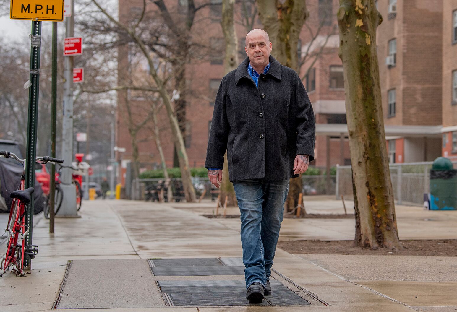 Bald man in his 40s in a dark gray coat and jeans walks on an urban sidewalk.