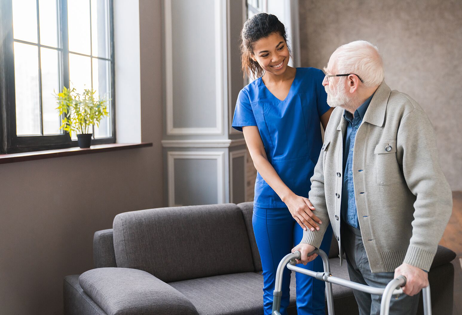 A female physician in blue scrubs walks to an elderly man who uses a walker.