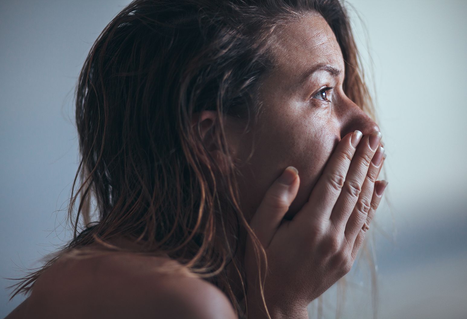A profile of a woman with wet, shoulder length hair. She looks up and away as she holds her hand to her face, covering her mouth.
