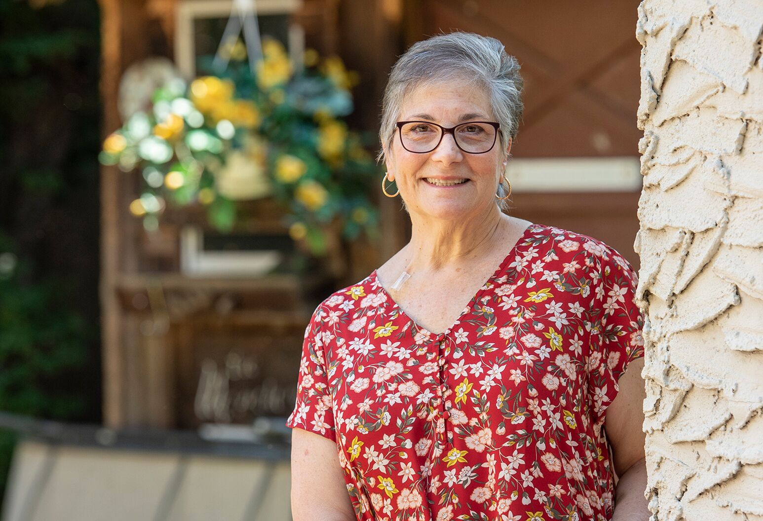 Woman with short, silver hair and red floral blouse smiles and leans against a wall outdoors.