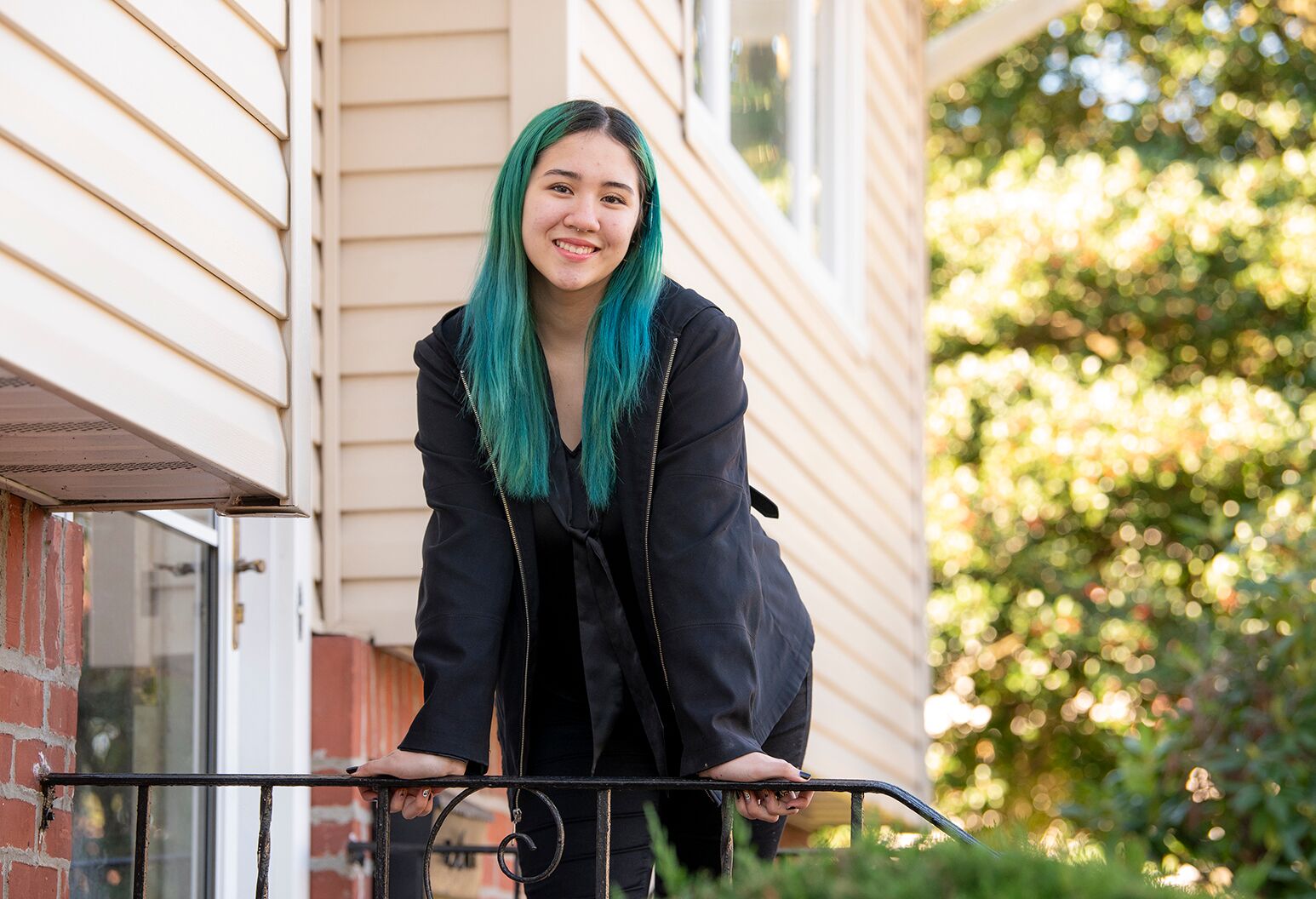 Young smiling woman with dyed green hair and black jacket leaning on a porch railing.