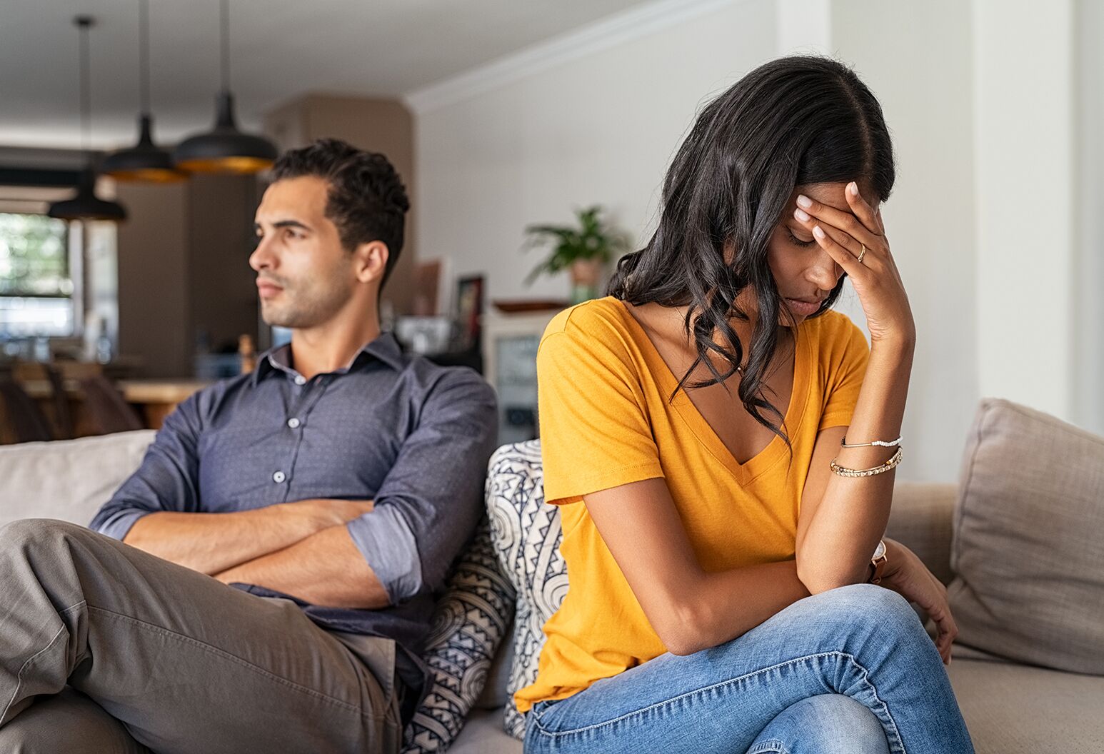 A couple sitting on the couch, with the woman looking upset and turned away from the man.