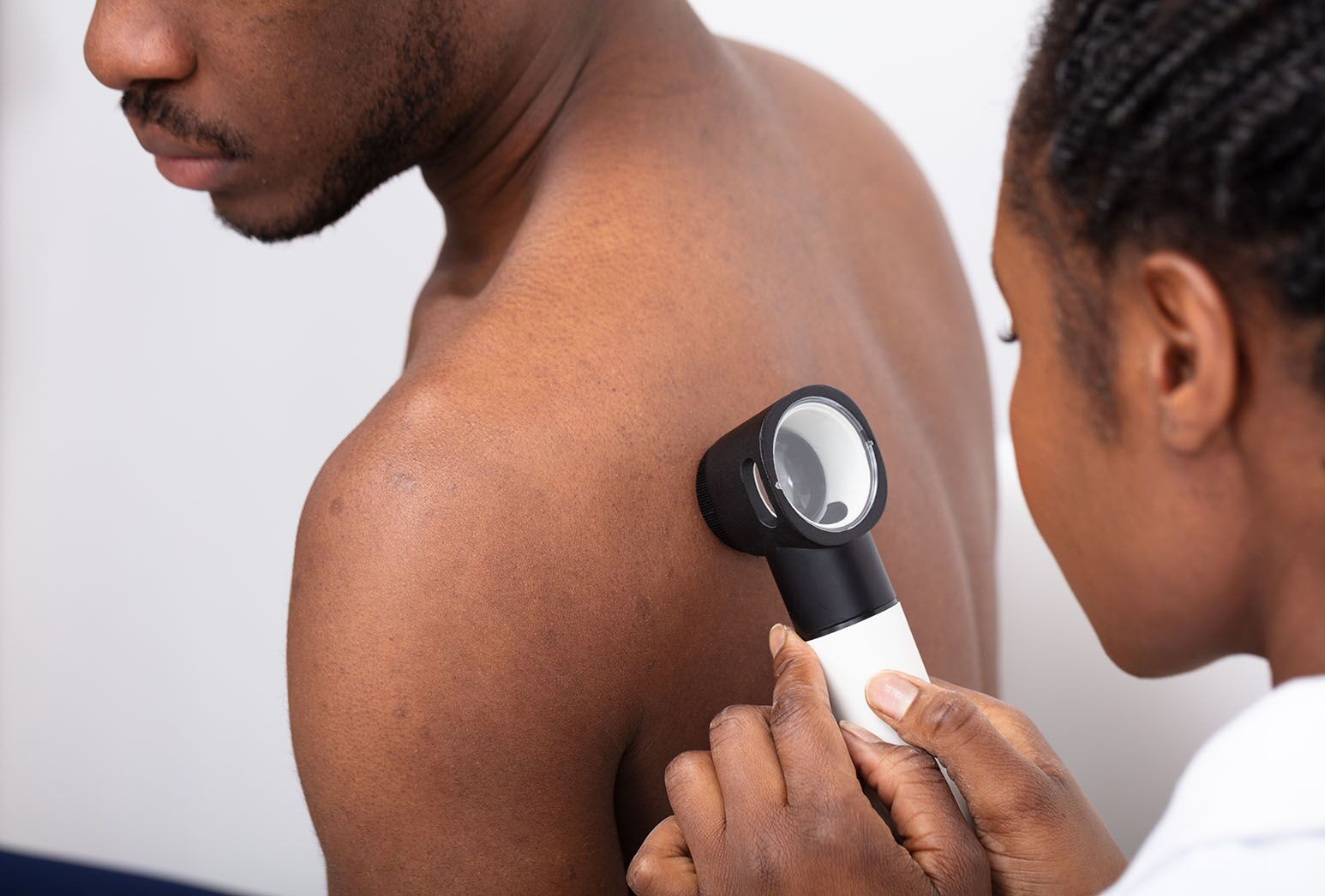 A dermatologist examines her patient's back using a dermatoscope; the patient, male, faces away.