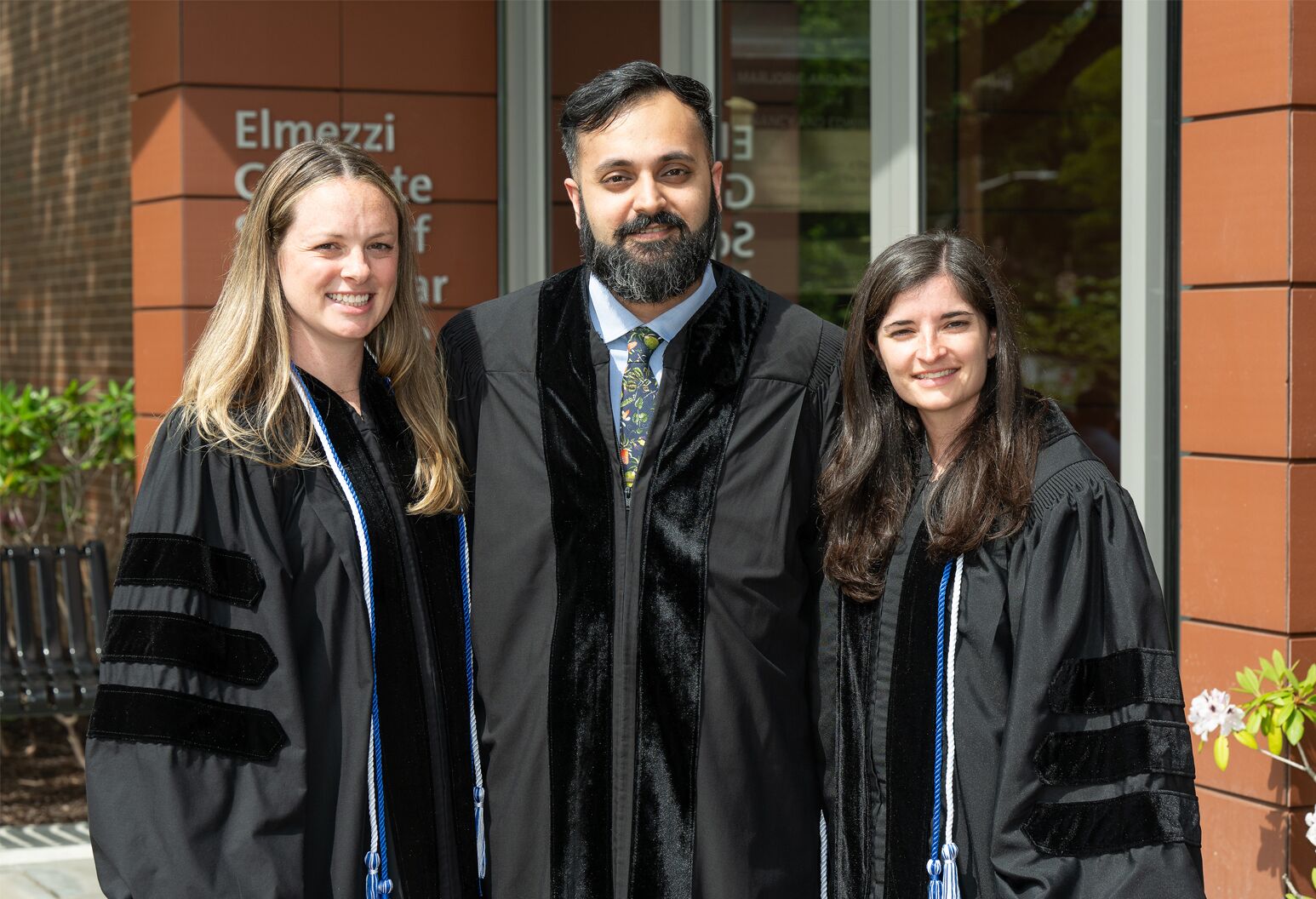 A man and two women stand side-by-side in dark graduation robes and cords and pose for a photograph.