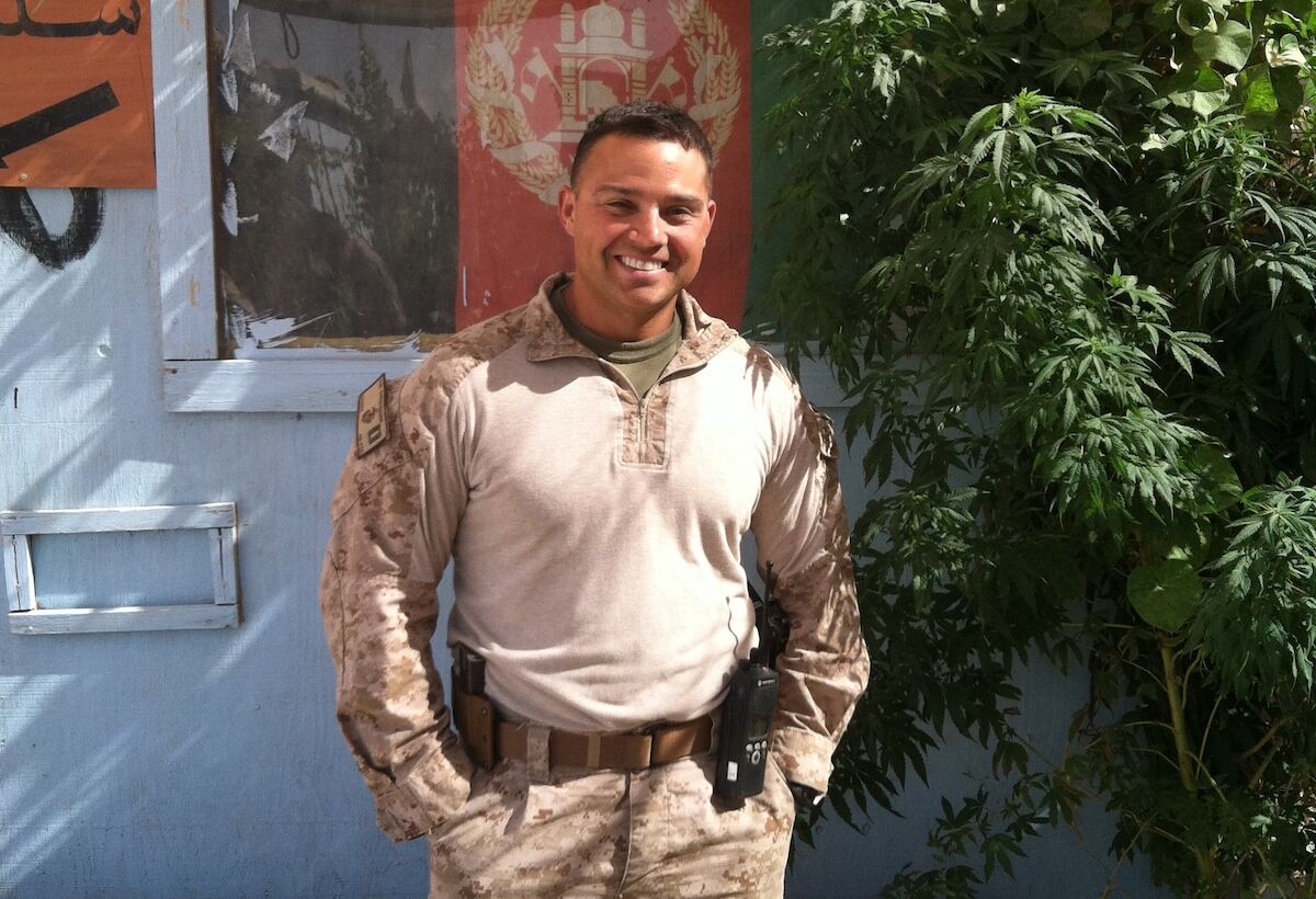 A man with short brown hair wearing military-style clothing stands outside in front of a tree and a blue building. 