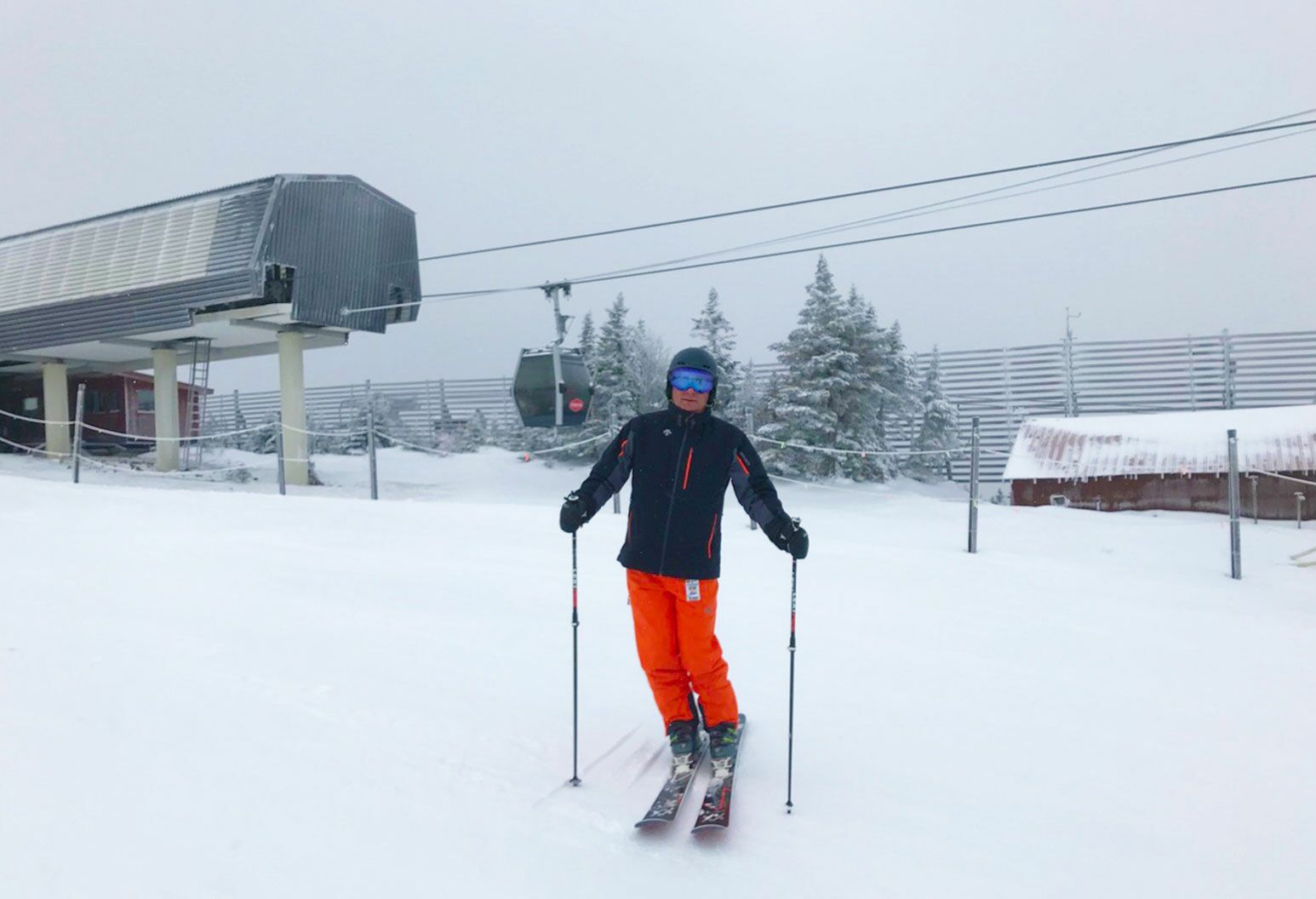 A man in red ski pants and black jacket wearing goggles poses for a camera while holding ski poles and skis in front of a ski-lift