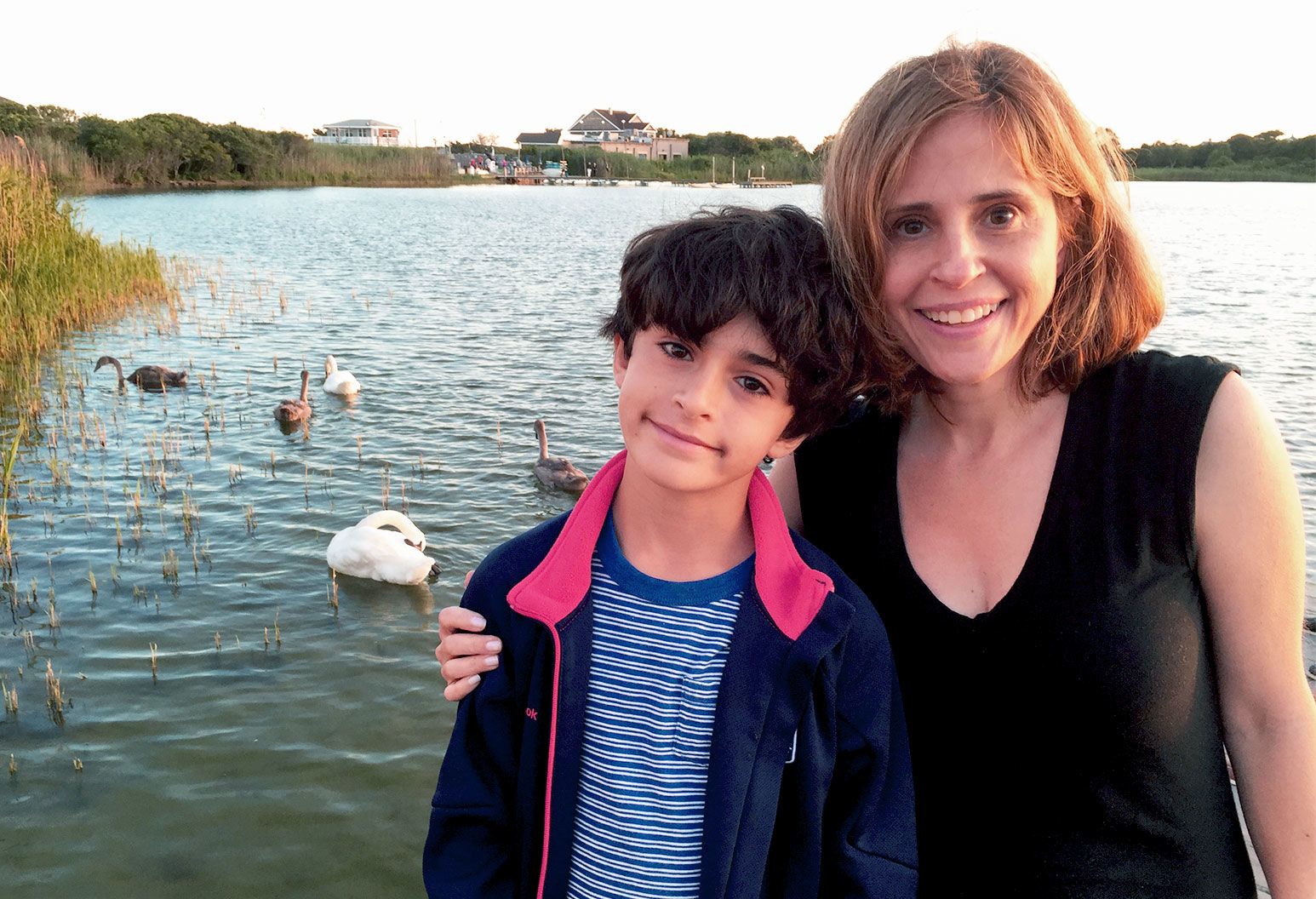 a young boy with curly black hair, a striped shirt displays a faint smile as he stands in front of a lake with an older woman with her hand on his shoulder. She has shoulder length auburn hair, and wears a black top. In the lake are geese treading in the water as the sun begins to set.