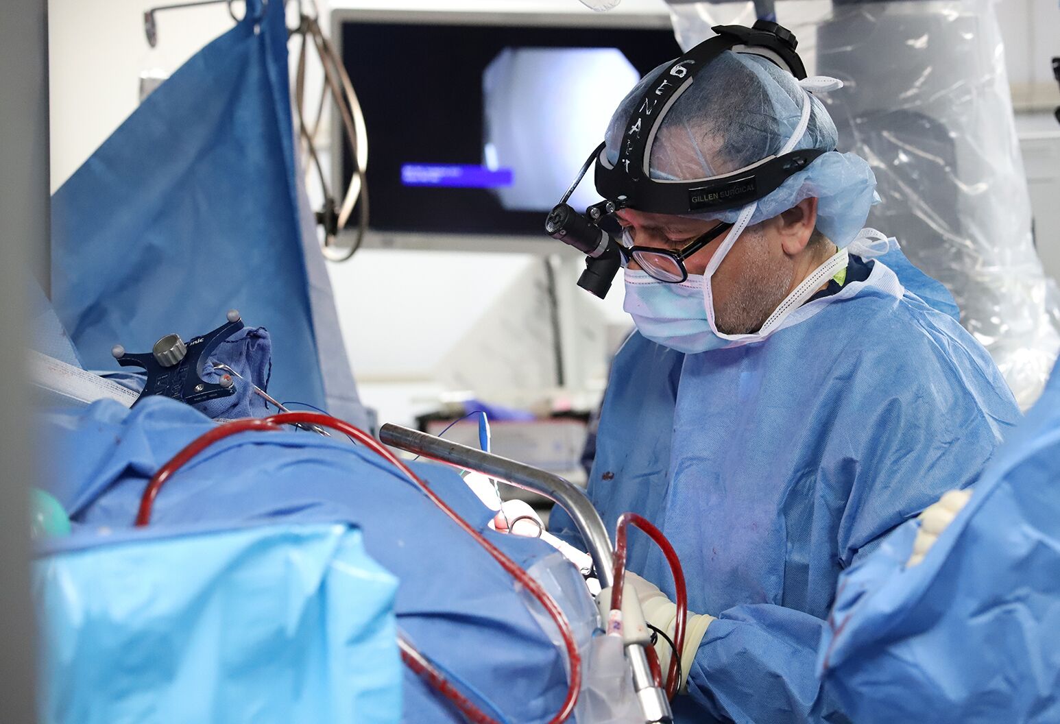 A male surgeon wearing scrubs and a headlamp operates on a patient's brain. 