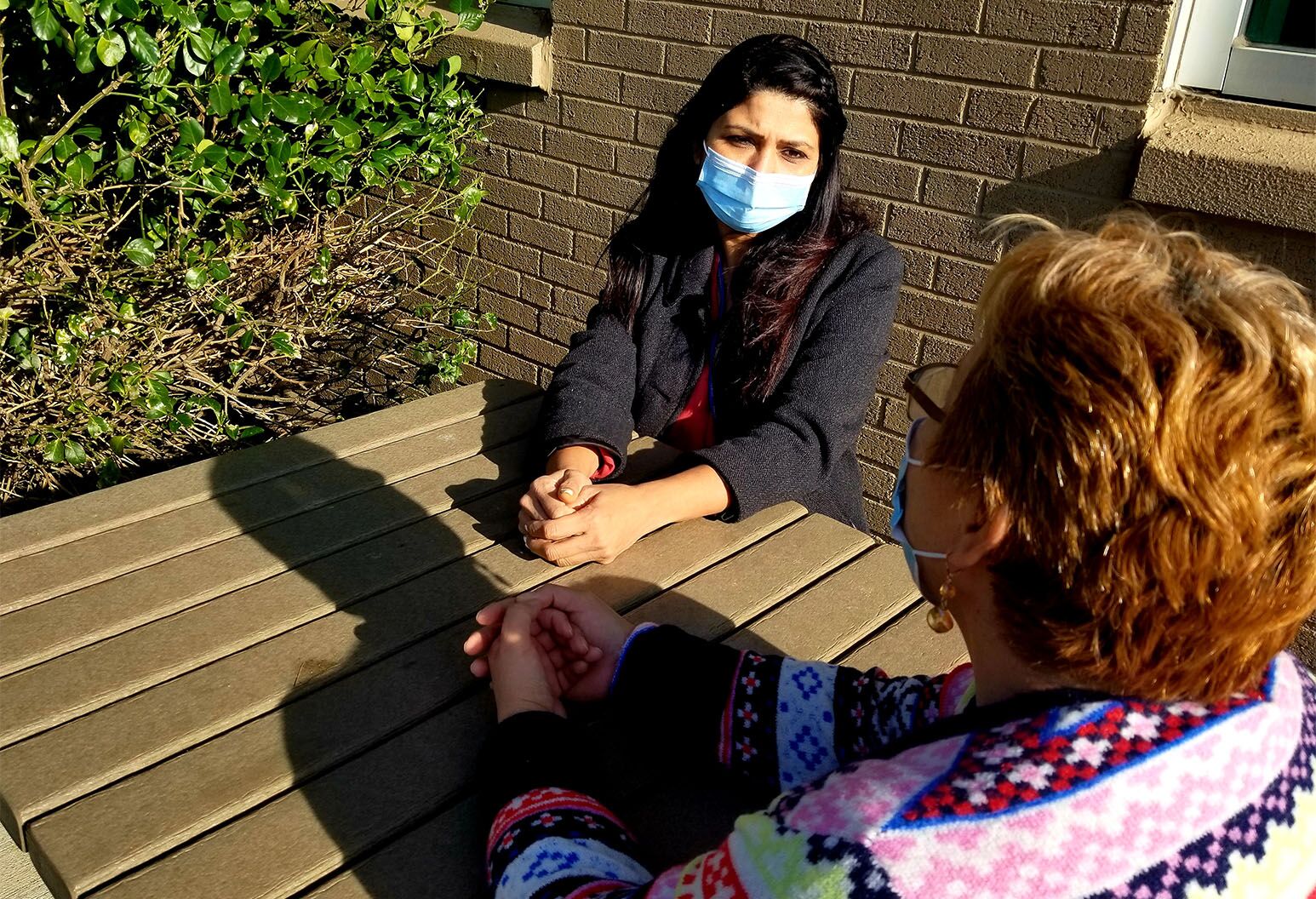 2 women wearing masks sit at a table outside.