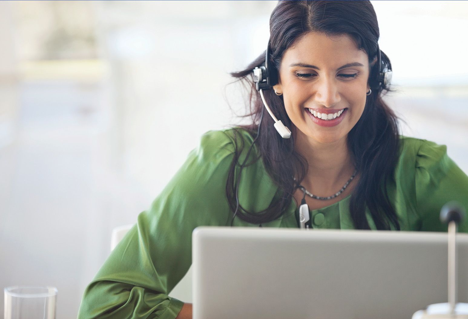 A woman in a green top wearing a headset smiles at the computer screen.