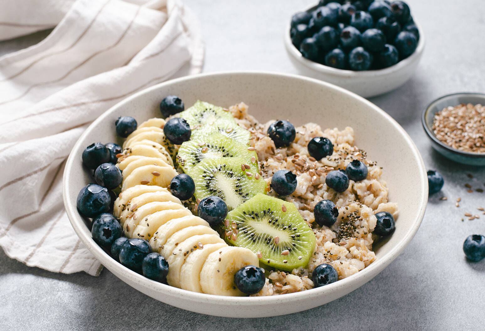 A bowl of oatmeal, kiwi, blueberries, and bananas, surrounded by a small bowl of grains and blueberries.