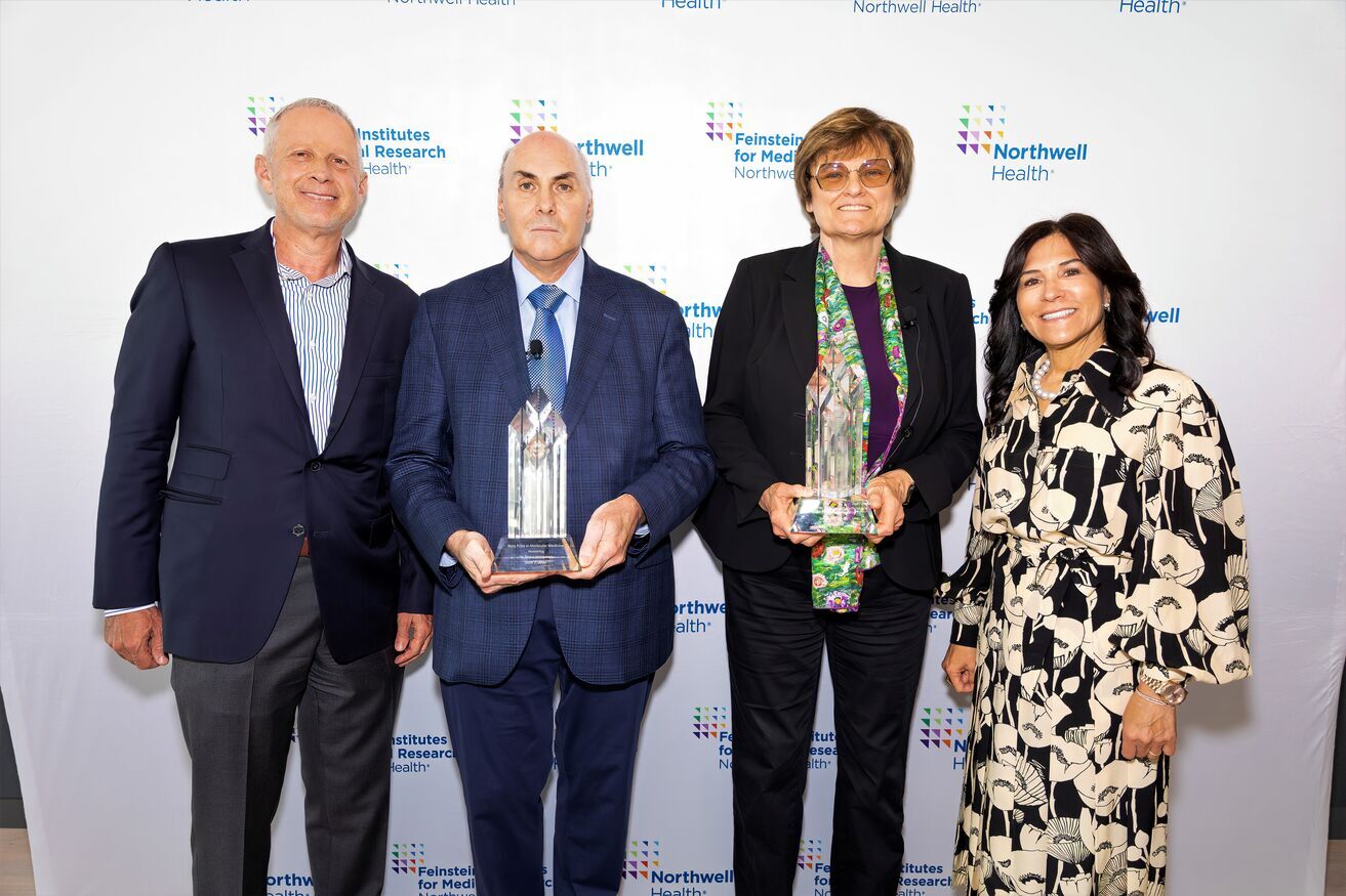 A man and woman stand against a Northwell backdrop. Each holds an award statuette.