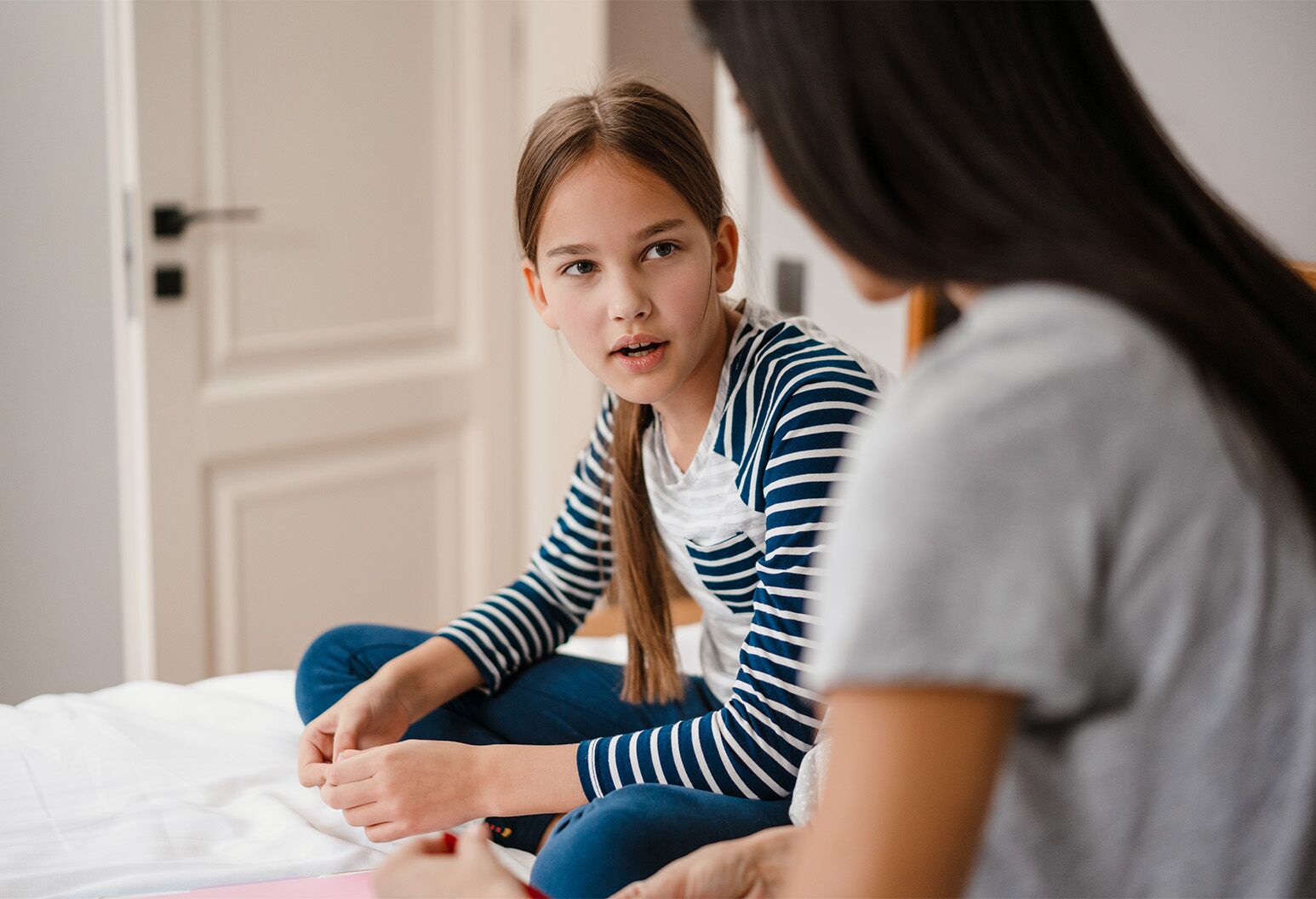 A young mother with long dark hair wearing a gray t-shirt speaks to her daughter, wearing a blue and white striped long-sleeve shirt, both sitting on a bed.