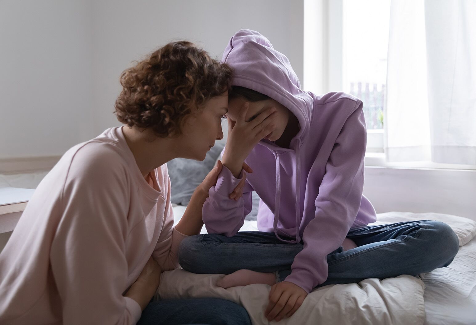 A mother with medium-length brown hair consoles a teenage girl wearing a pinkish-purple hoodie who is sitting on a bed with her hand over her face.