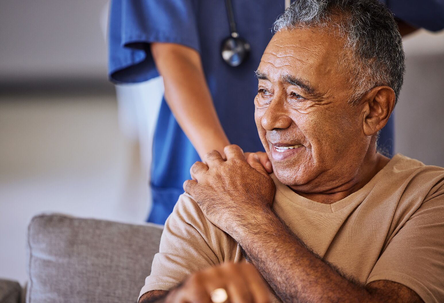 Older man with brown and gray hair sits on couch as female medical professional stands behind him with her hand on his shoulder