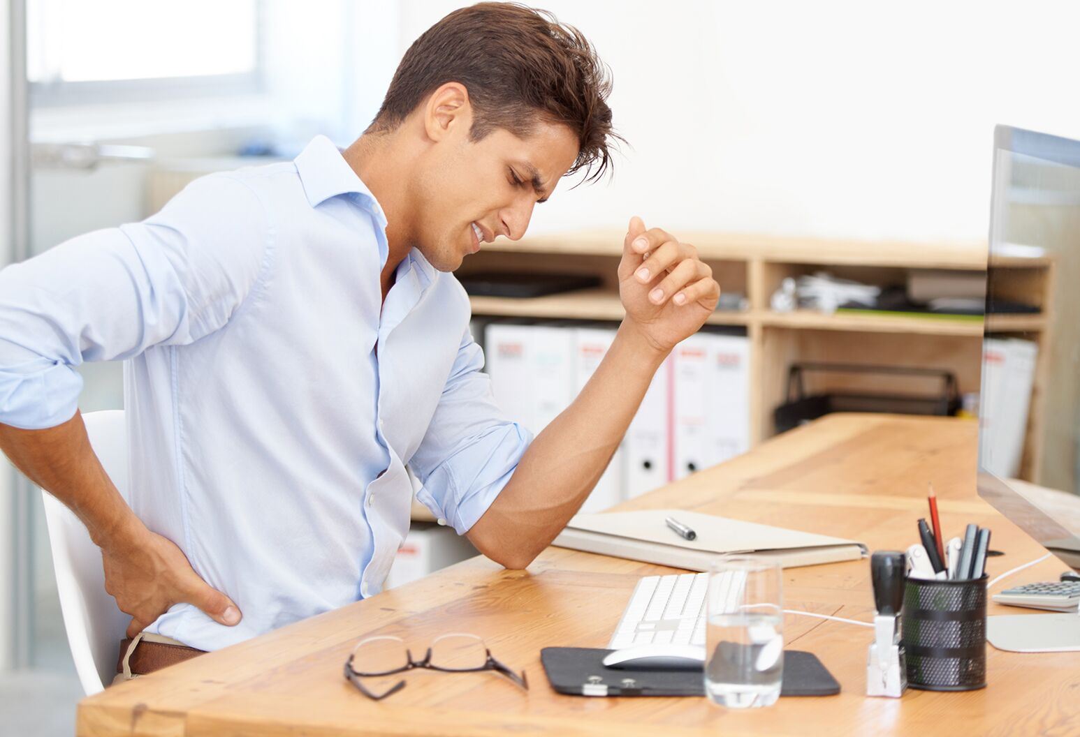 businessman rubbing his aching back while sitting at his desk in front of his computer 