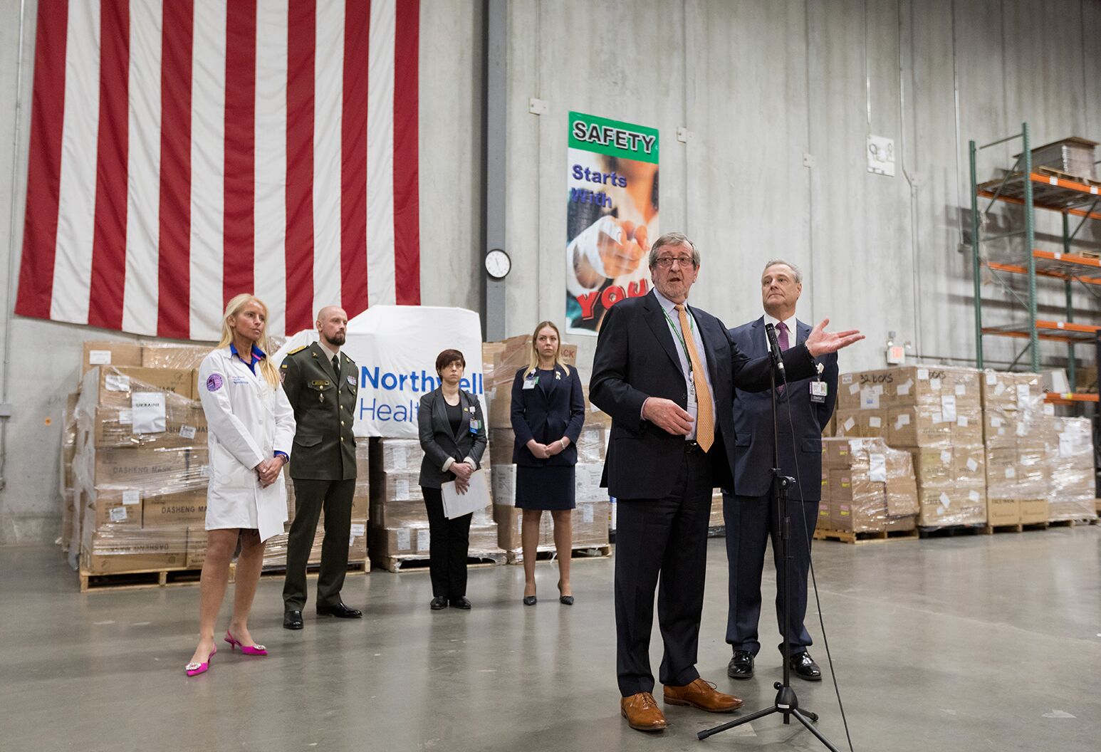 Man standing in a room speaking with people and a large stack of boxes in the background