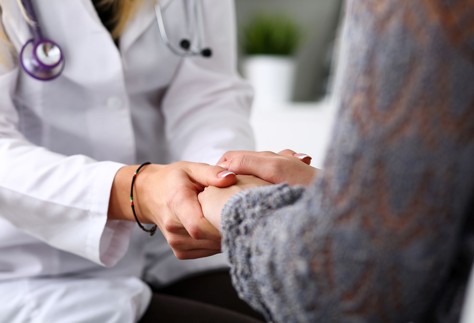 With only hands visible, female doctor with white lab coat and stethoscope is shown holding hands with female patient.